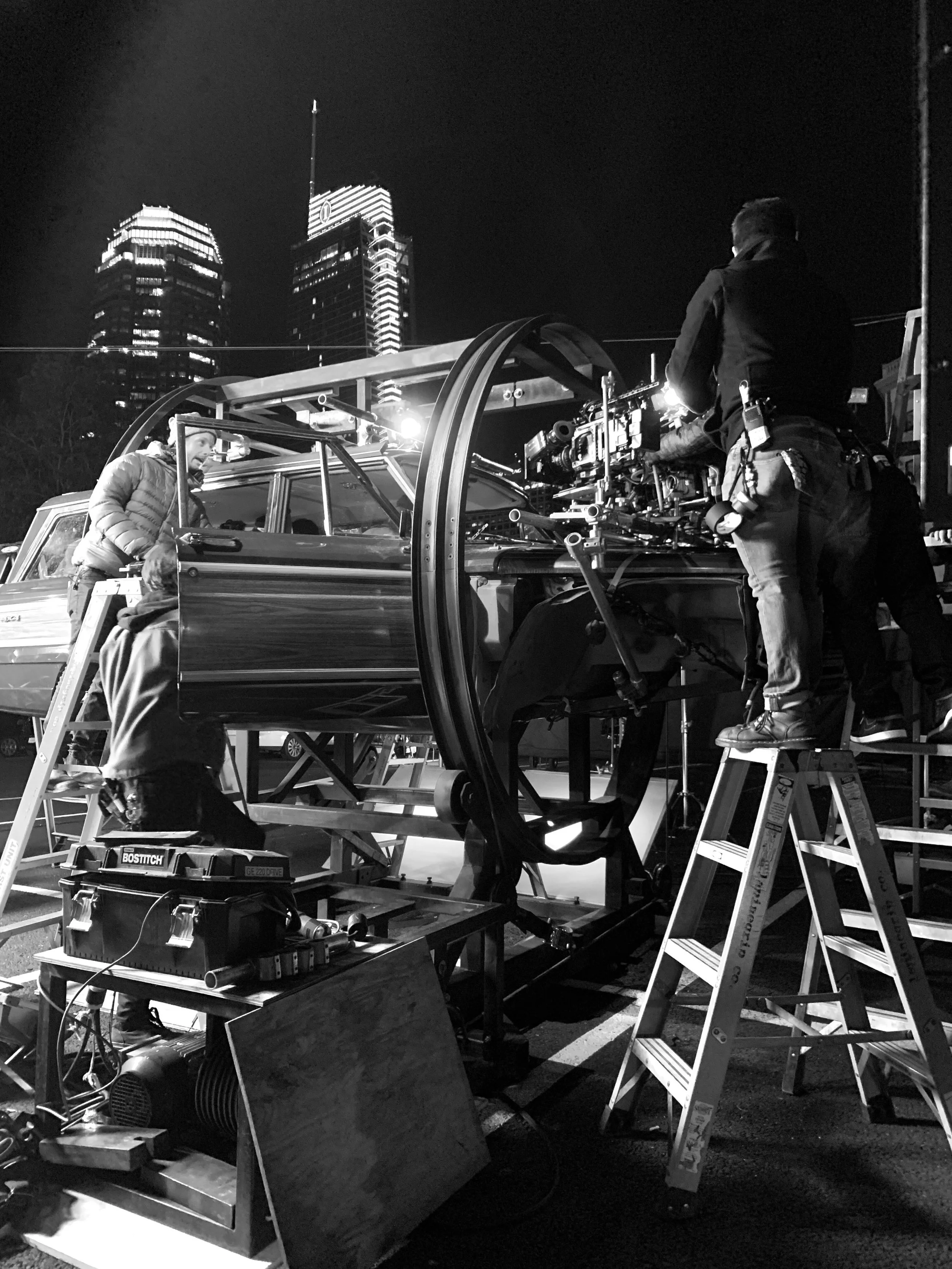 Nighttime behind-the-scenes shot of a film or television production crew setting up camera equipment on a vehicle with city skyscrapers in the background.