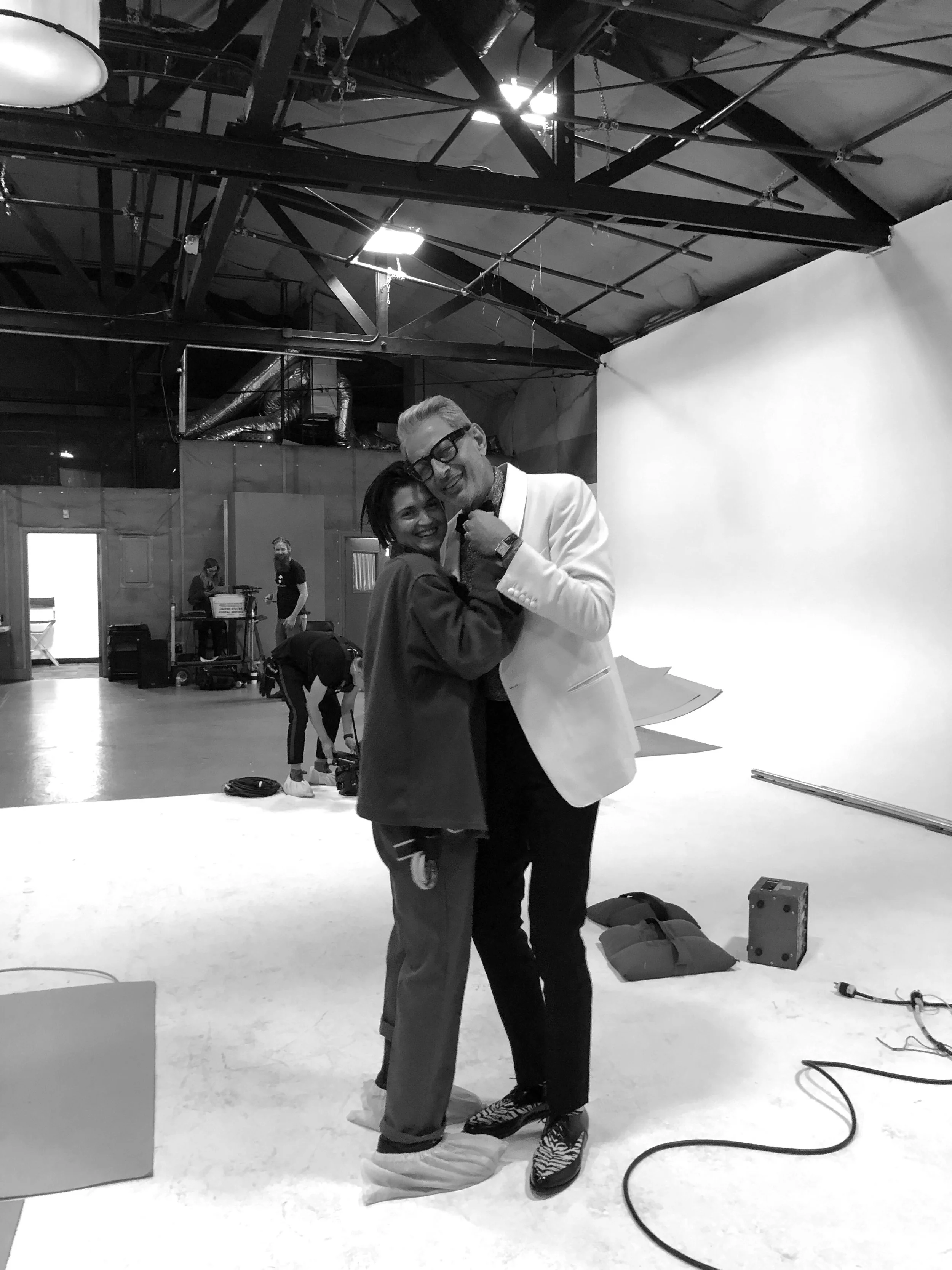 Two people smiling and hugging each other in a photography studio with a plain white background. The man is wearing a white suit with glasses, and the woman is dressed casually. There are studio lights, equipment, and crew members in the background.