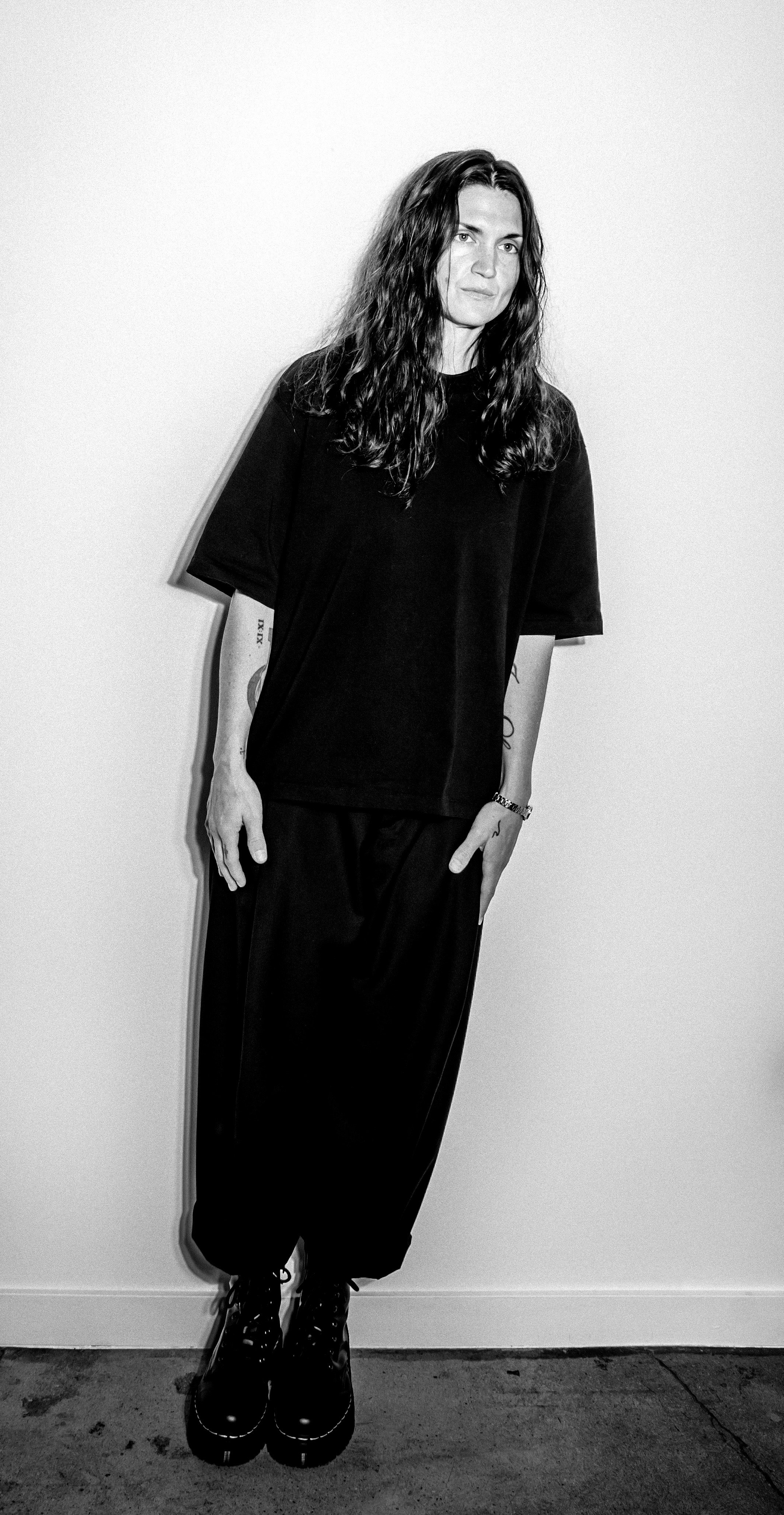 Black and white photo of a woman with long wavy hair wearing a loose black T-shirt, black pants, and black combat boots, standing against a plain white wall.