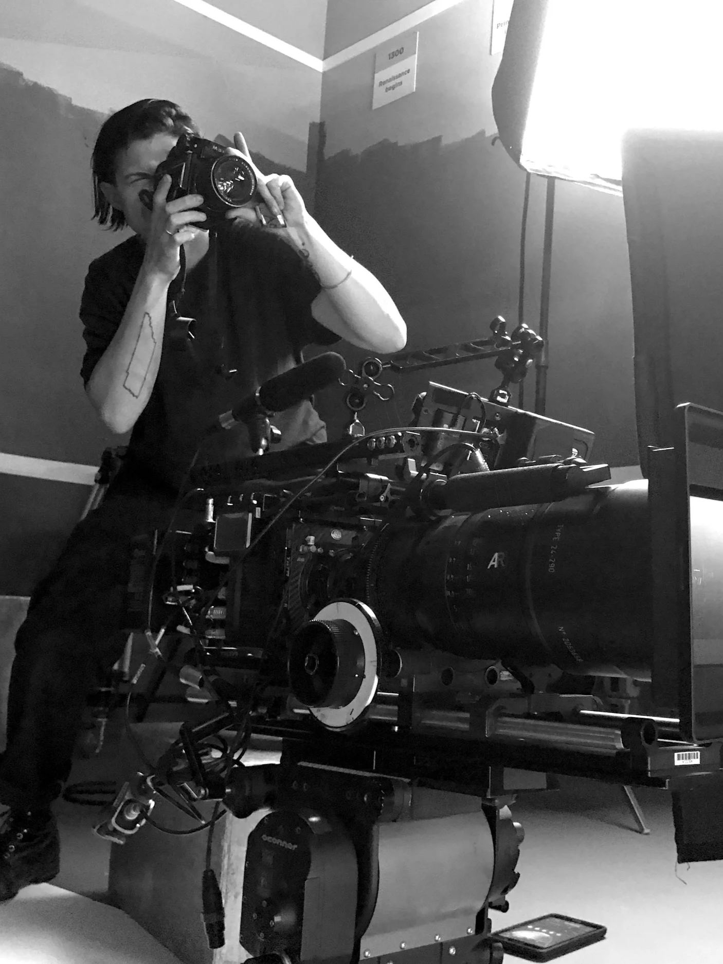 A person taking a photograph in a mirror using a professional camera, surrounded by camera equipment and a light source in a room.