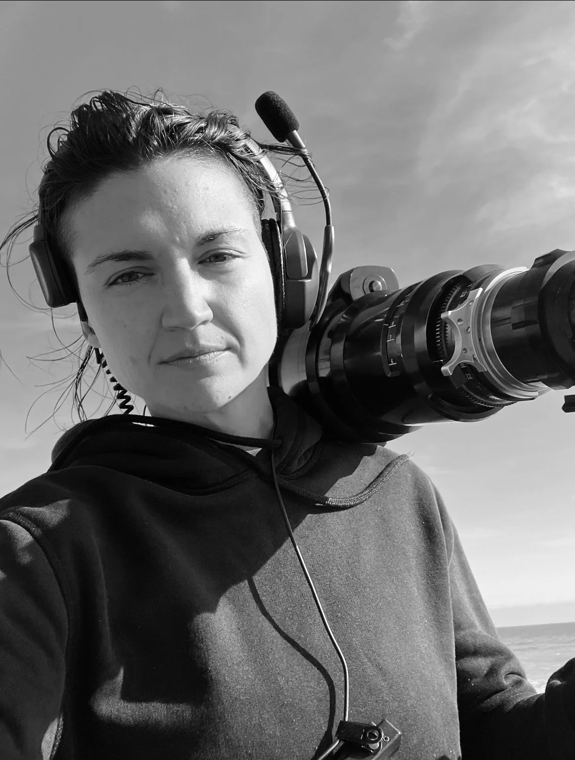 Black and white photo of a woman wearing a headset and holding a camera with a large lens. She is outdoors, with a cloudy sky in the background.
