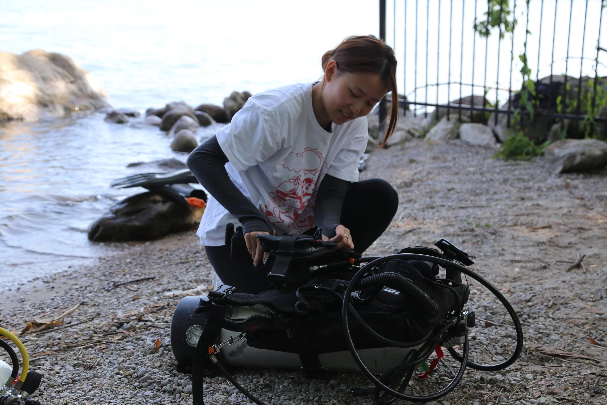 A woman with short hair, wearing a white t-shirt and black long-sleeve shirt, bending down over a black bicycle near a rocky lake shore, with a metal fence and water in the background.