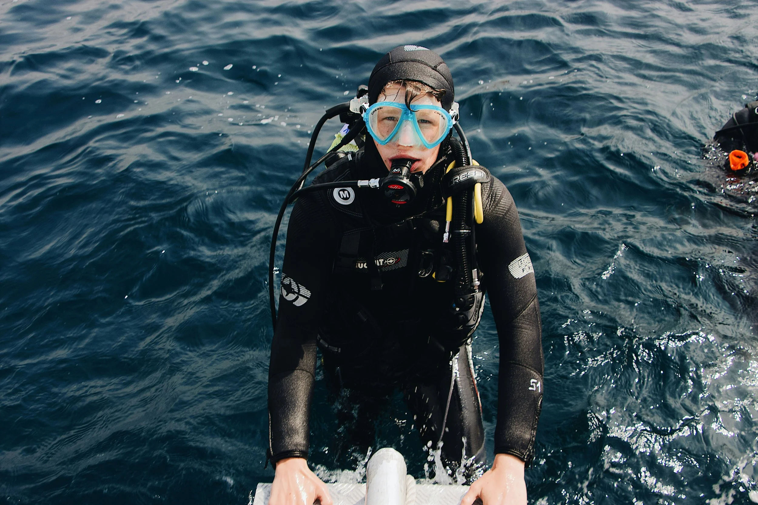 A scuba diver in a wetsuit and diving mask emerging from the water, holding onto a boat's edge, with the ocean surface surrounding him.