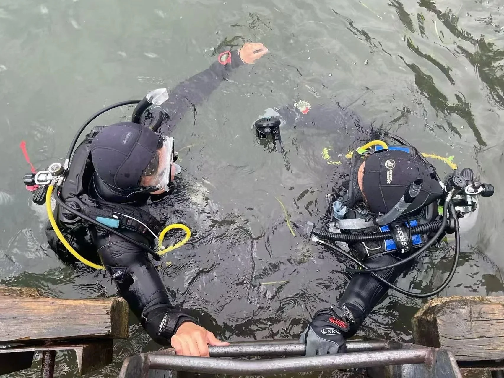 Two scuba divers in full gear are in a body of water, one is in the water while the other is at the water's edge holding onto a ladder.