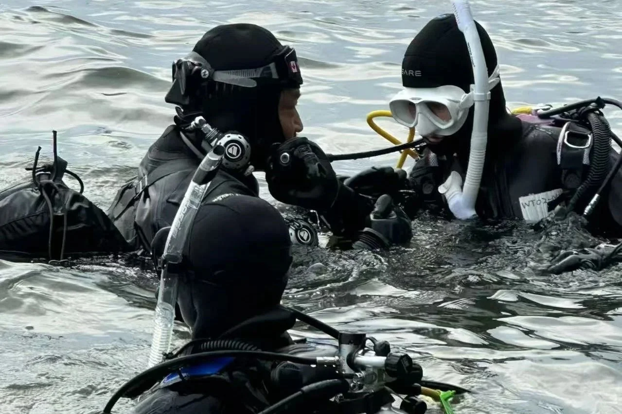 Two scuba divers in black wetsuits and masks underwater, one assisting the other with equipment, in a body of water.