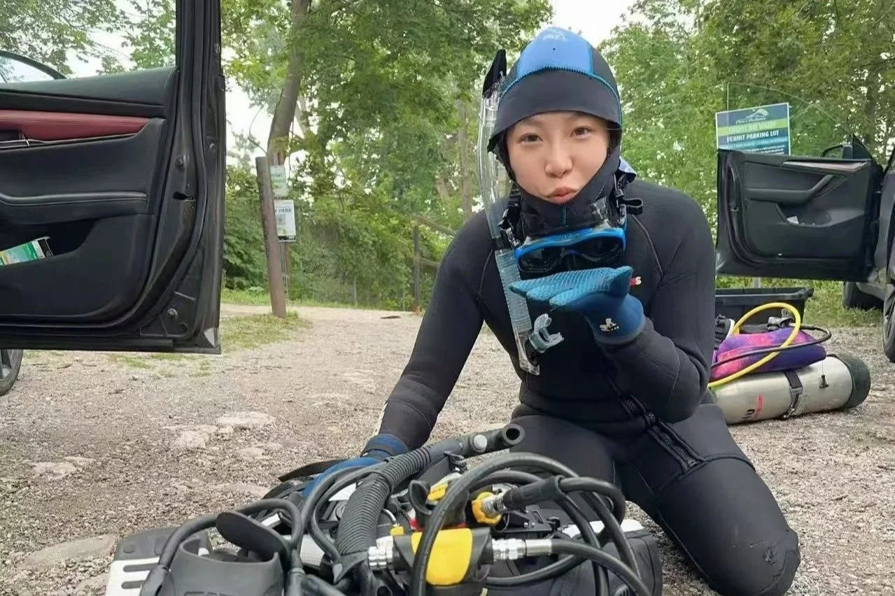 A person dressed in black gear and a helmet kneeling on gravel ground, holding a diving mask and a glove, surrounded by open car doors and scuba diving equipment, in a wooded outdoor setting.