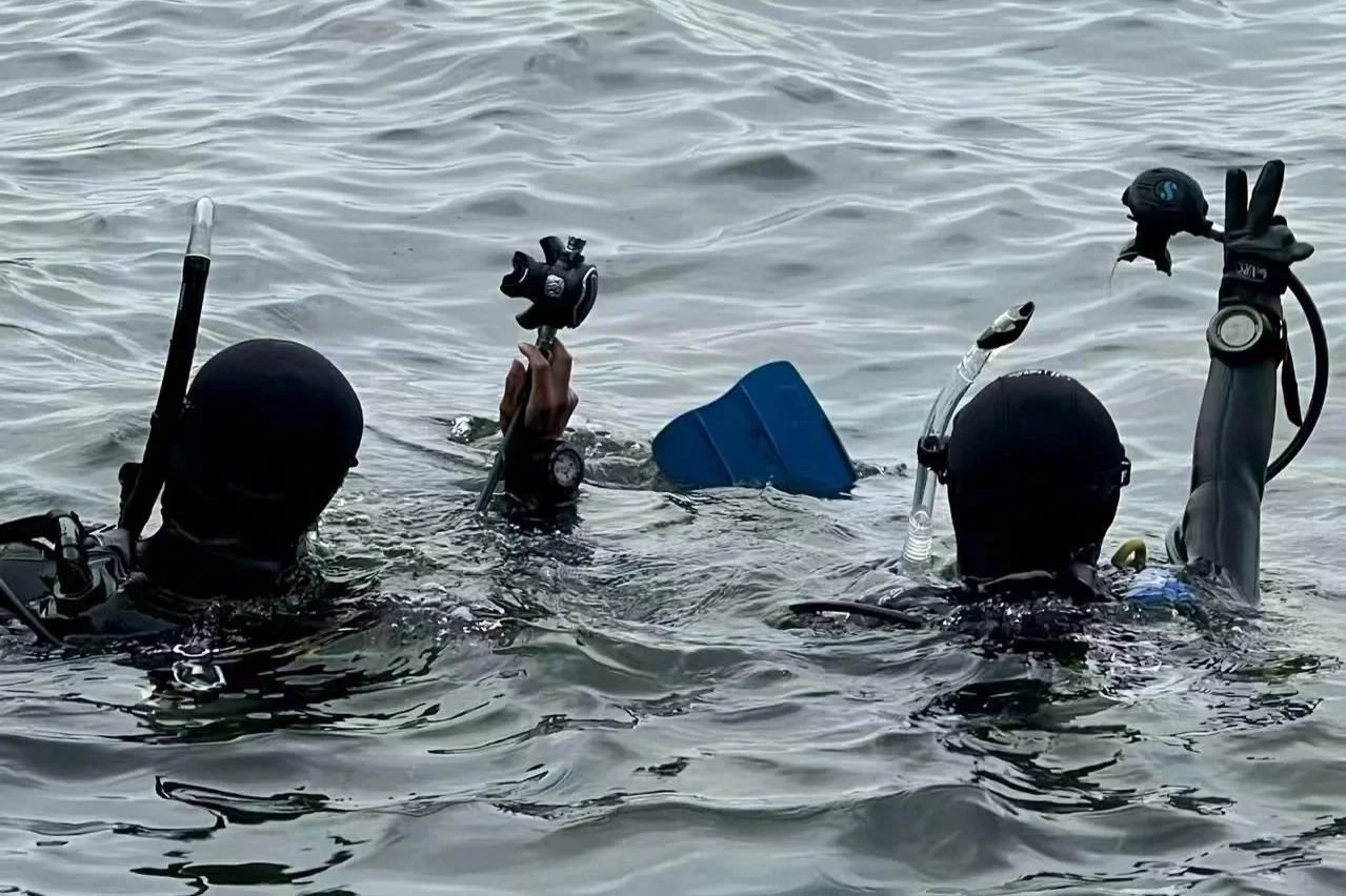 Two scuba divers holding underwater gear in the water, with one holding a camera and the other holding a snorkel, on a body of water.