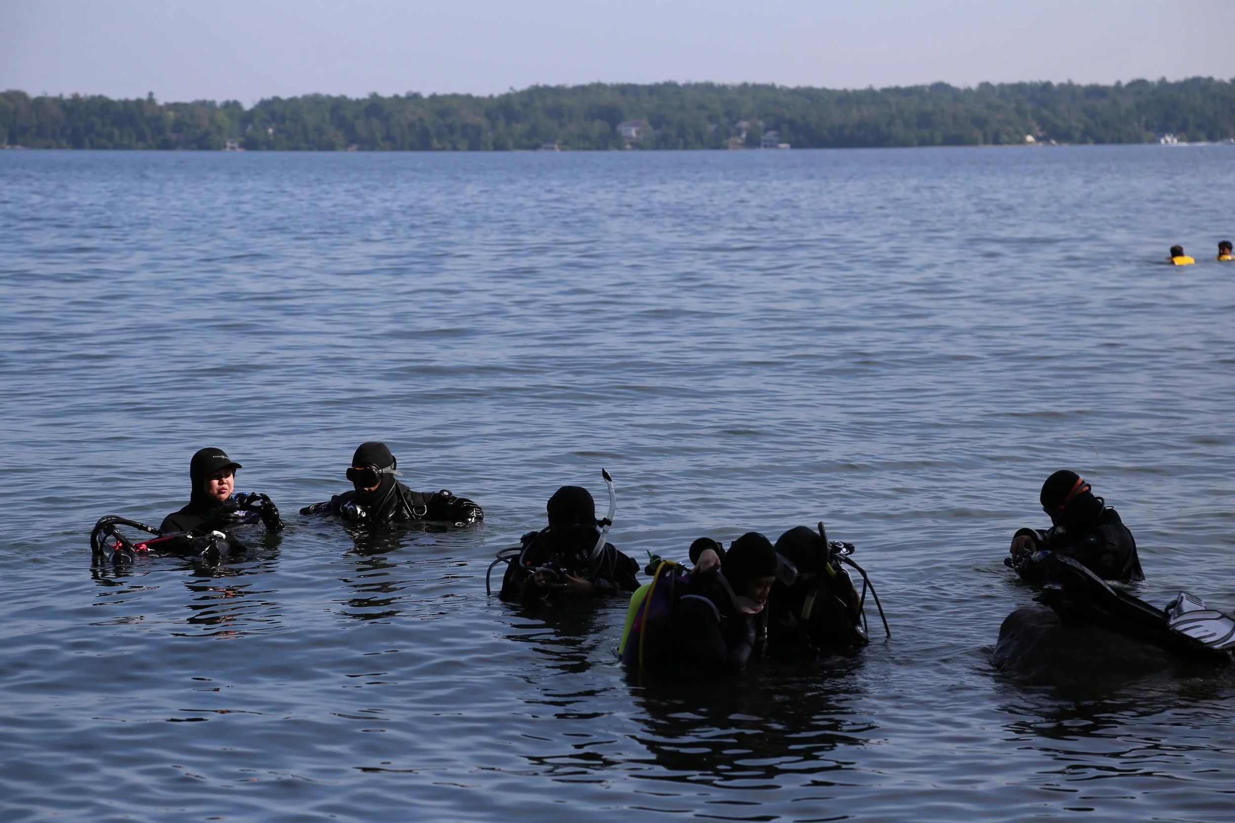 Group of people in scuba diving gear sitting and standing in a body of water, with two people swimming in the background.