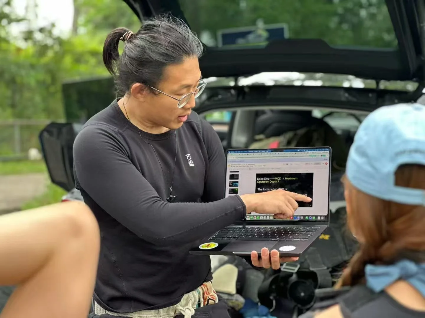 Woman with glasses showing a laptop screen to another person with a blue cap, outdoors near an open car trunk.