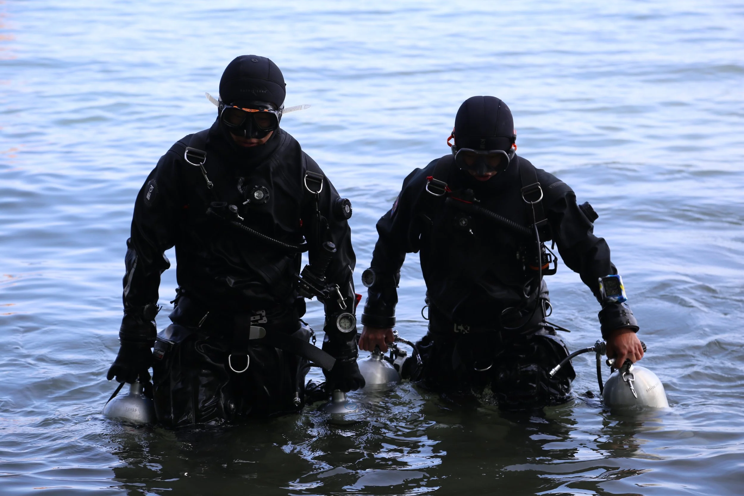 Two scuba divers in black wetsuits and diving gear standing in shallow water at the shoreline, preparing for a dive.
