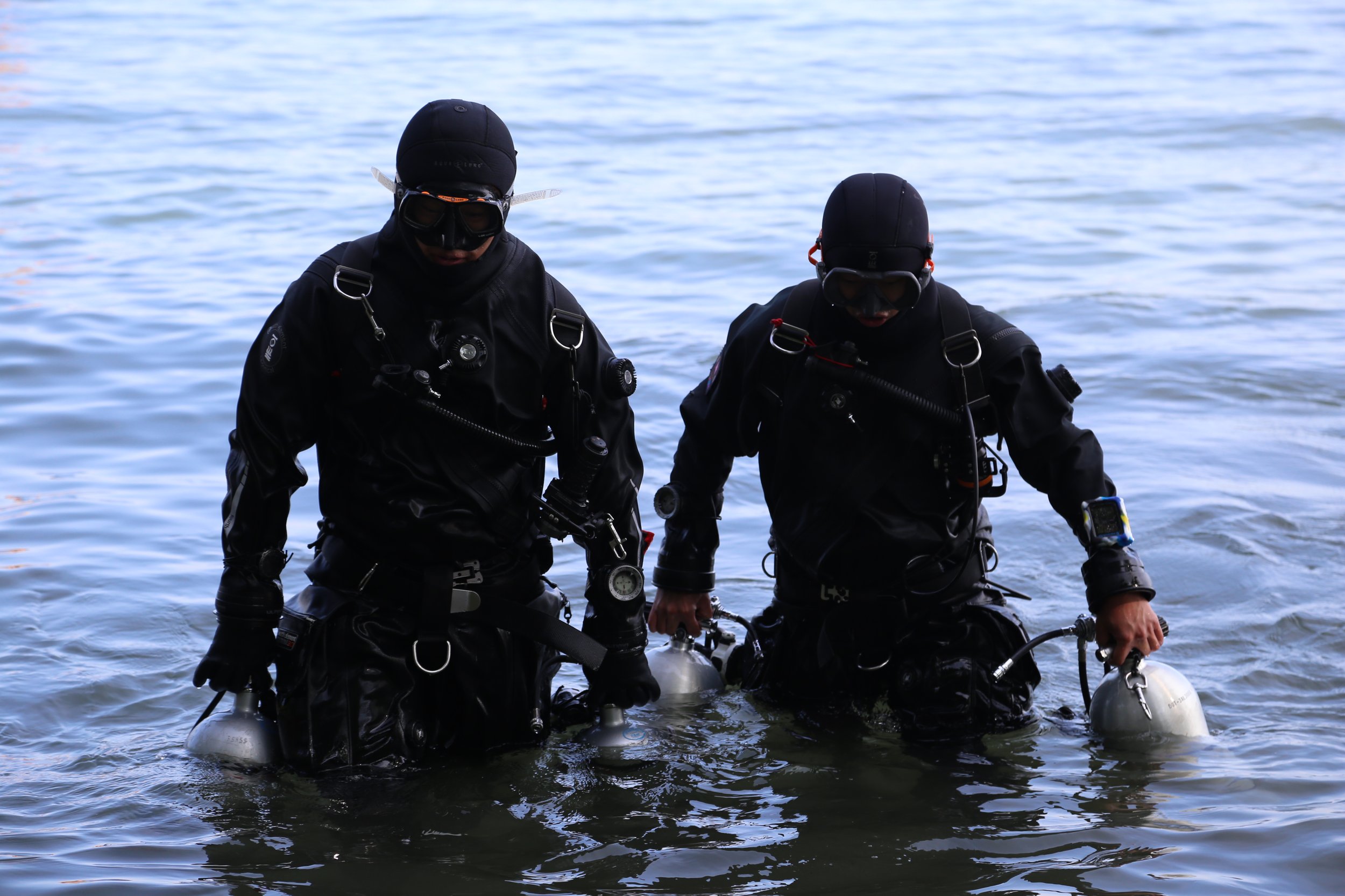 Two scuba divers in black wetsuits and helmets standing in shallow water near a lake, preparing for a dive.