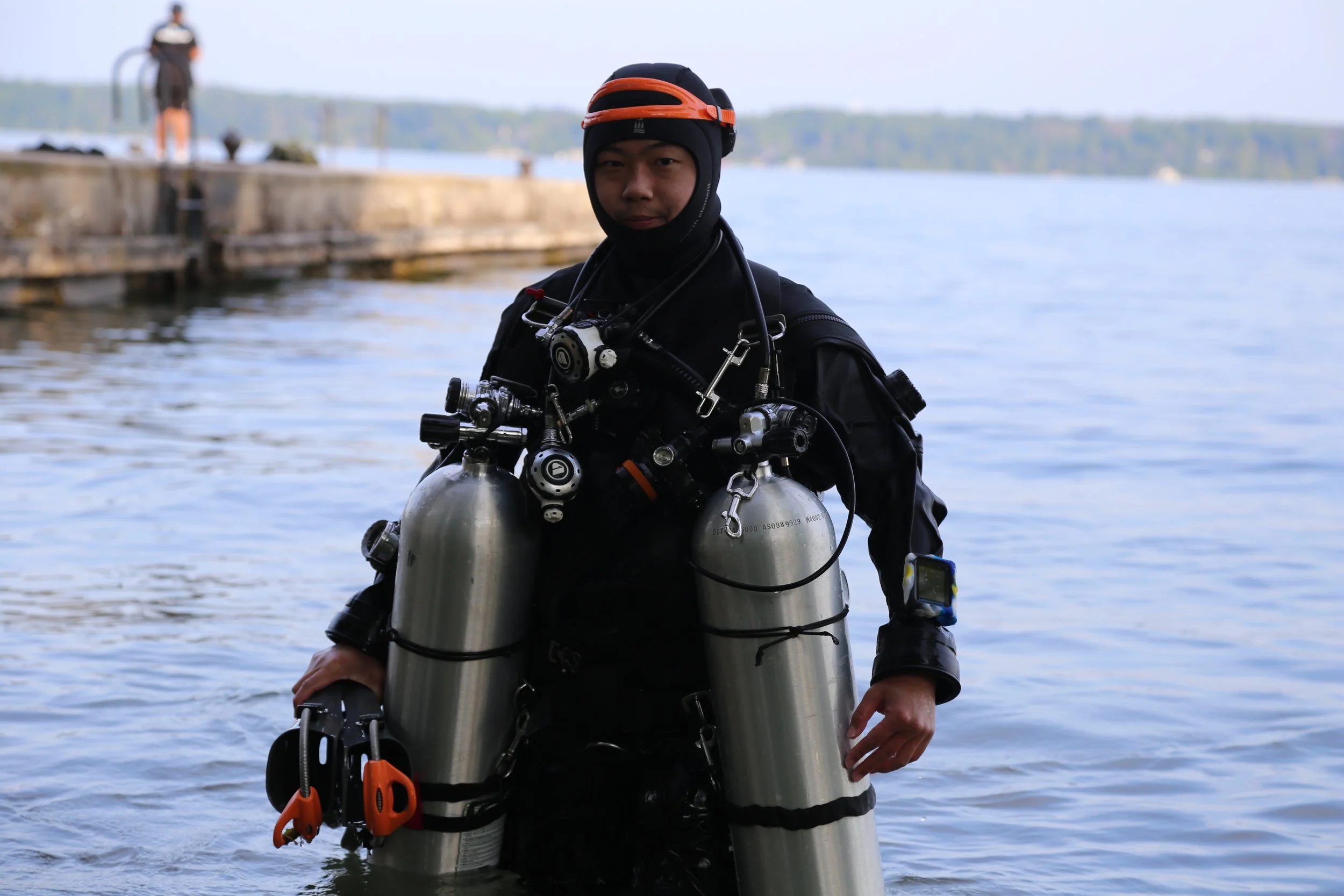 A person in scuba diving gear standing in water near a dock, with another person on the dock in the background.