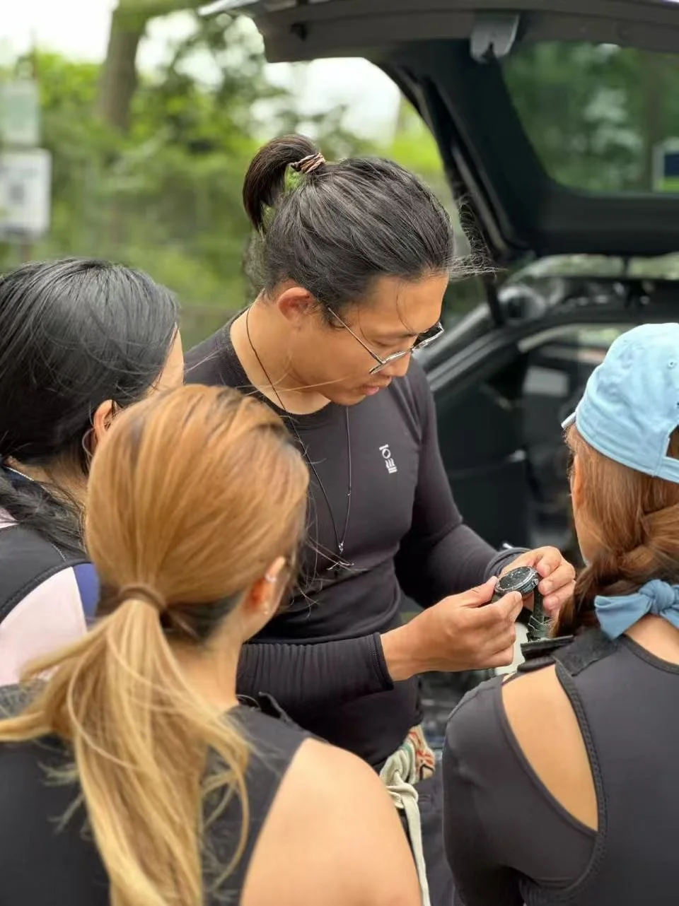 A woman with glasses and dark hair tied back shows a watch to three other women, all dressed in athletic clothing, near an open car trunk outdoors with trees in the background.