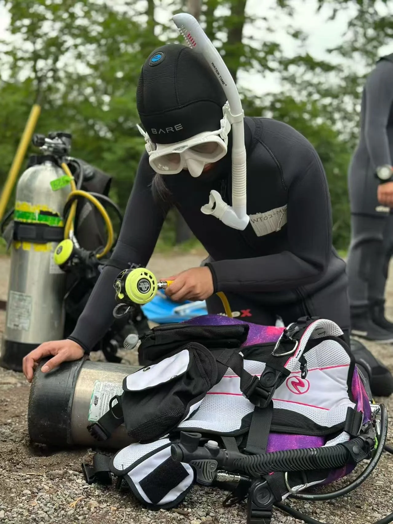 A person in diving gear, including a wetsuit, mask, and snorkel, adjusting equipment next to scuba tanks and a backpack on a gravel surface outdoors.