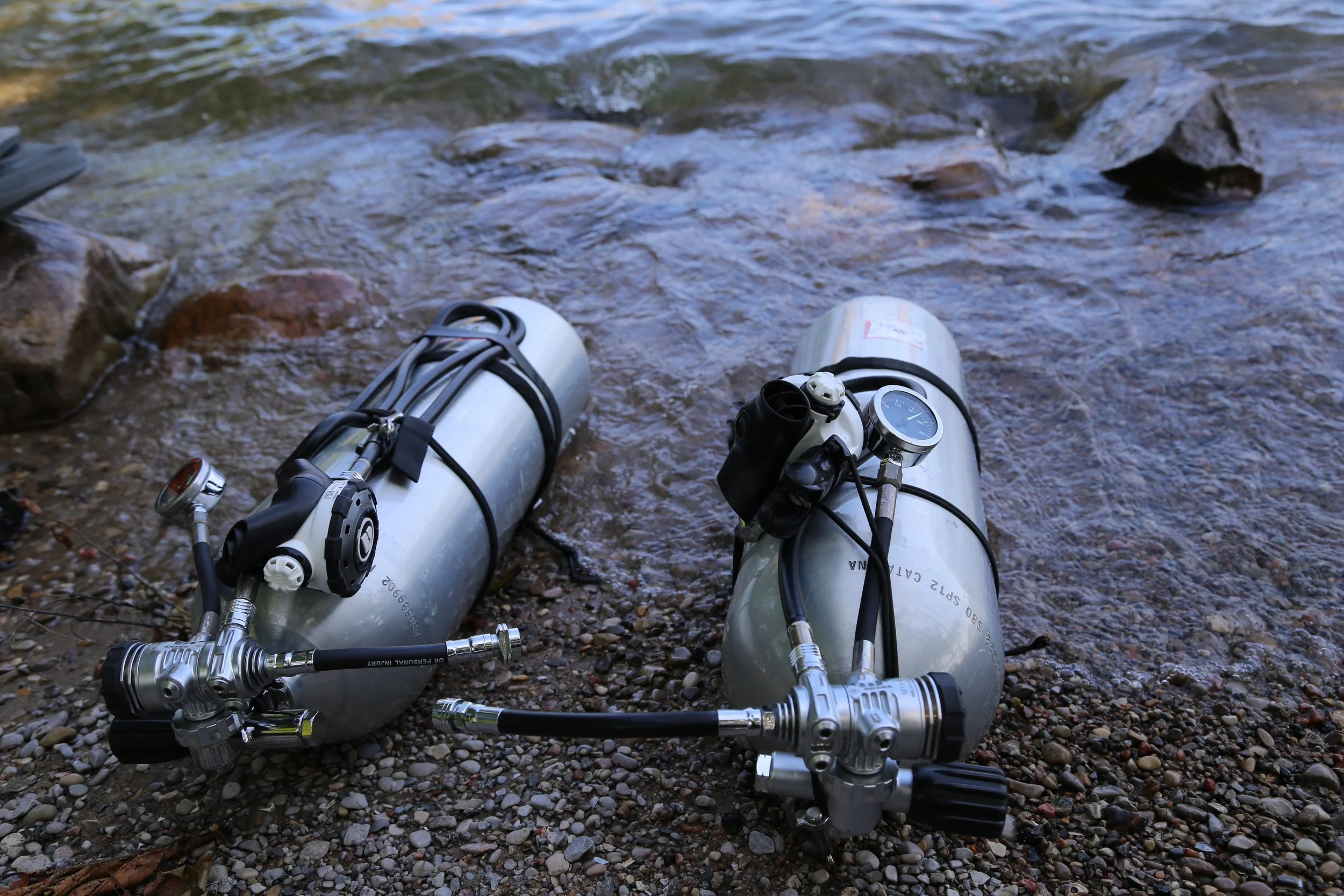 Two scuba diving tanks with regulators and gauges on the rocky shore of a river or lake, with water flowing nearby.