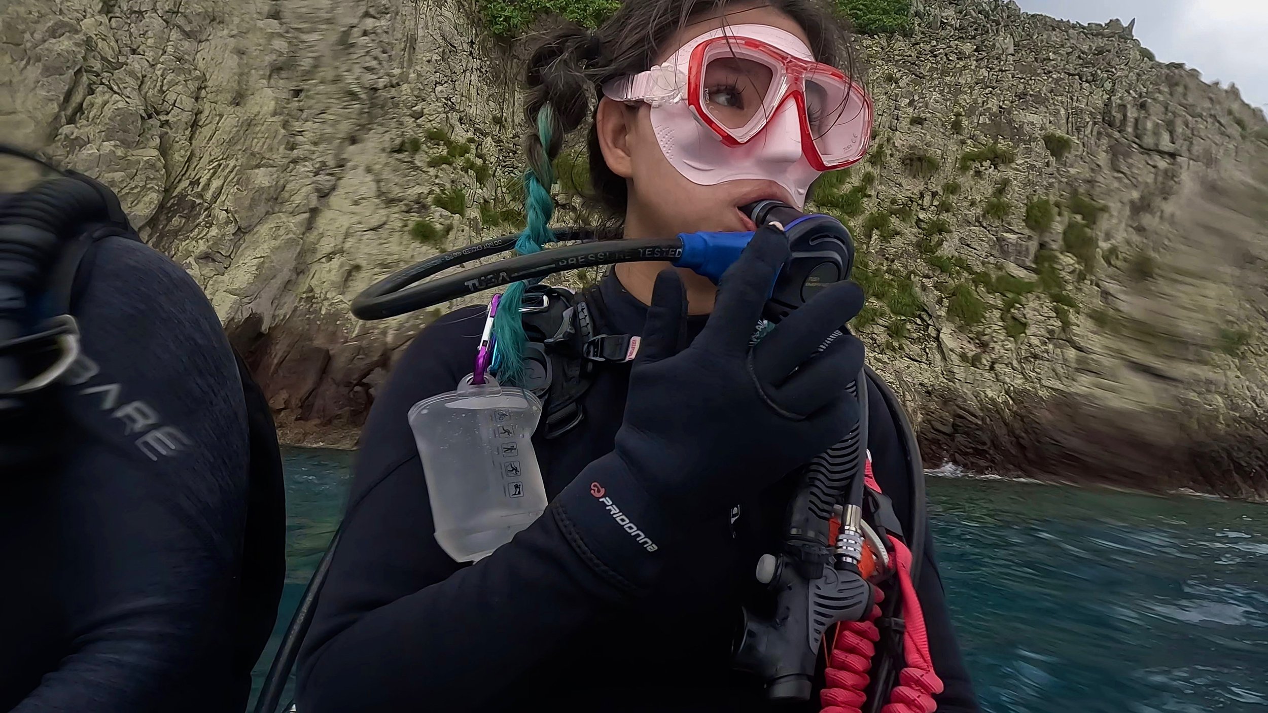 A woman wearing a pink mask with red goggles and black gloves on a boat, surrounded by water and steep rocky cliffs with greenery.