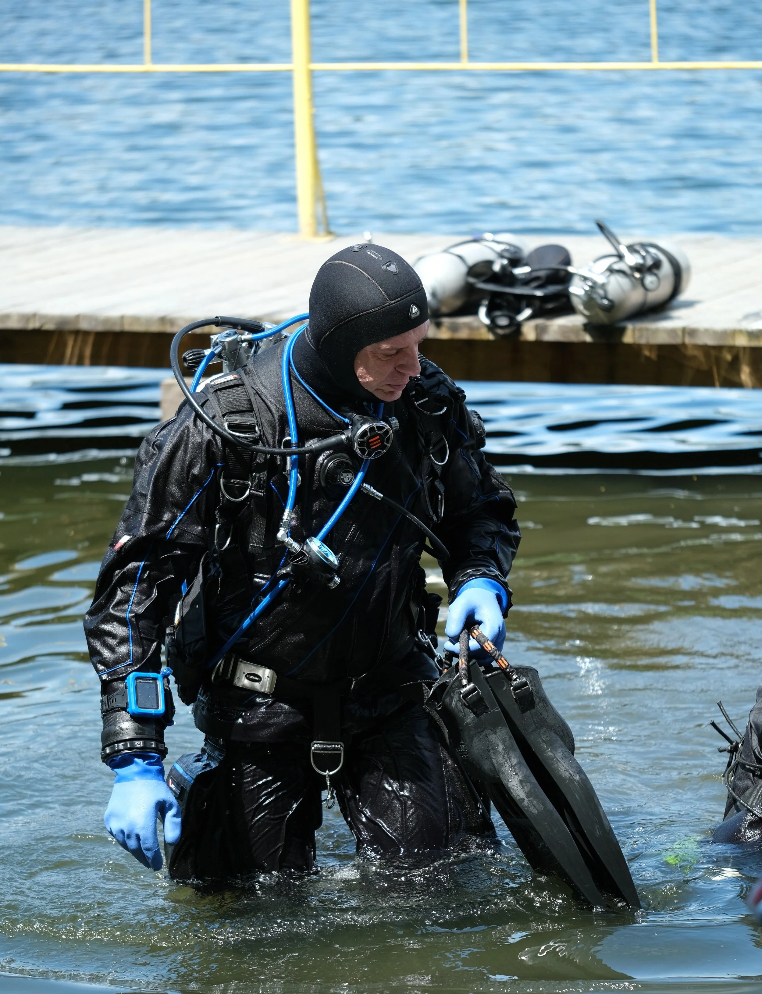 A scuba diver in a wet suit standing in water near a dock with scuba gear and fins visible.