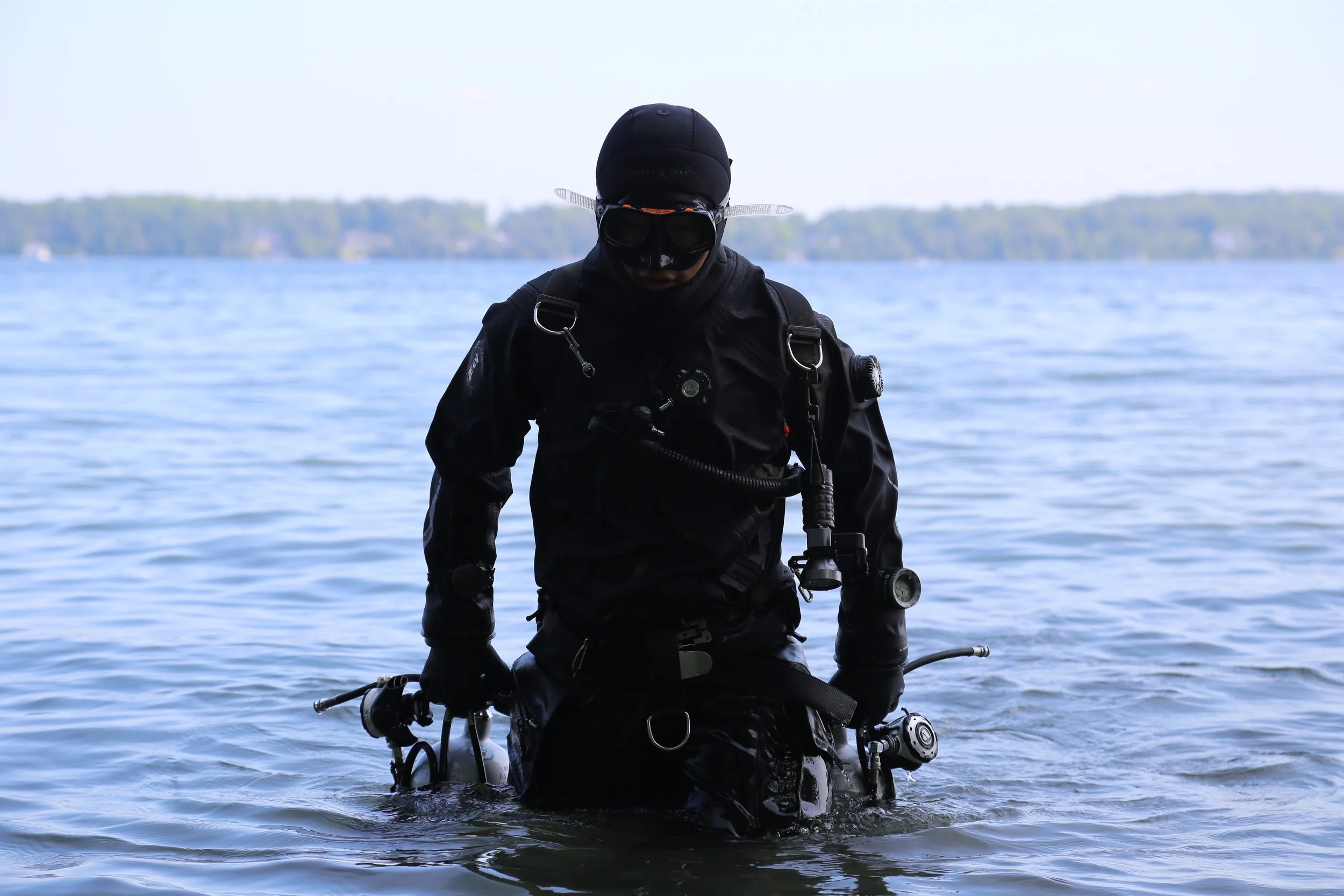 A diver dressed in black scuba gear, including a wetsuit, mask, and gloves, stands in shallow water near a shoreline with trees and hills in the background.