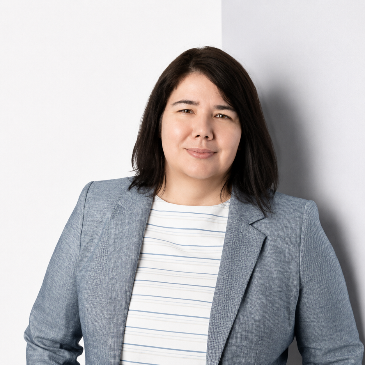 A woman with dark brown hair wearing a light gray blazer and a white striped shirt standing against a white background.