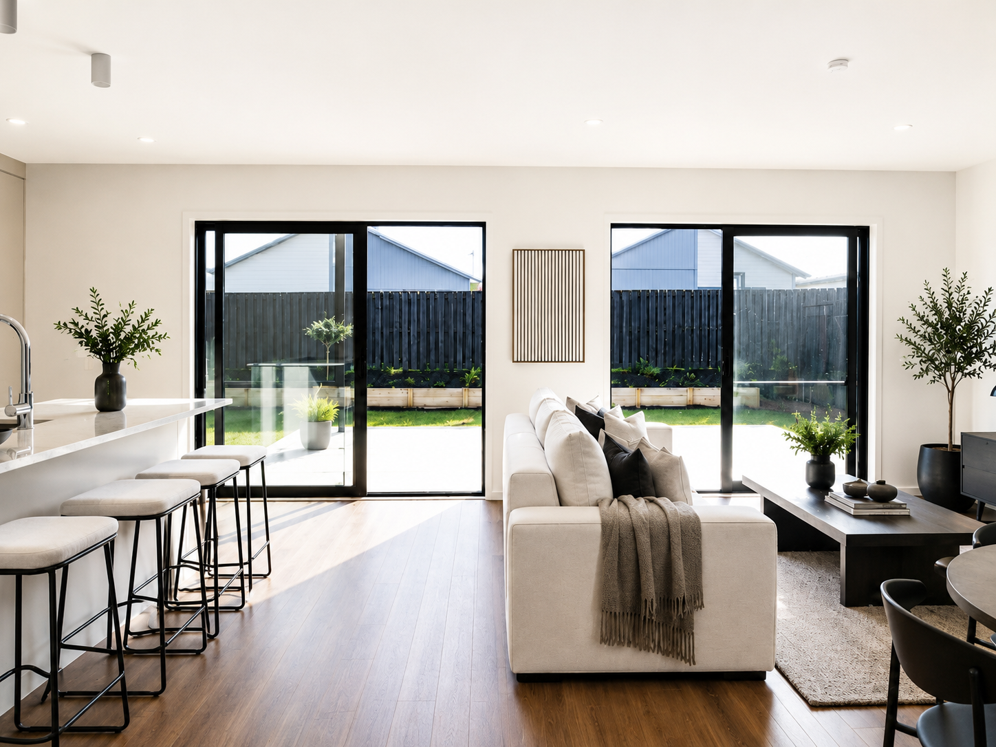 Modern living room with large sliding glass doors leading to a backyard, featuring a white sofa with pillows and a throw blanket, a black coffee table with decorative items, and green plants in black pots.