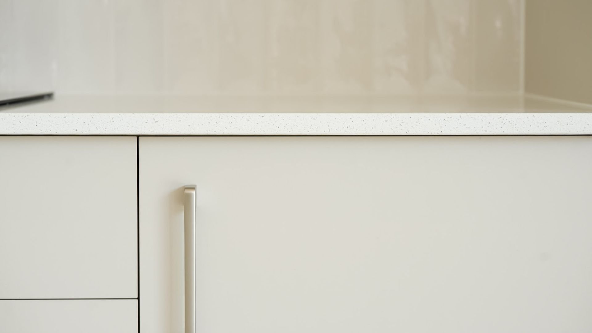 Close-up of a beige kitchen cabinet with a silver handle, topped with a speckled white kitchen countertop. Background shows beige tiled wall.