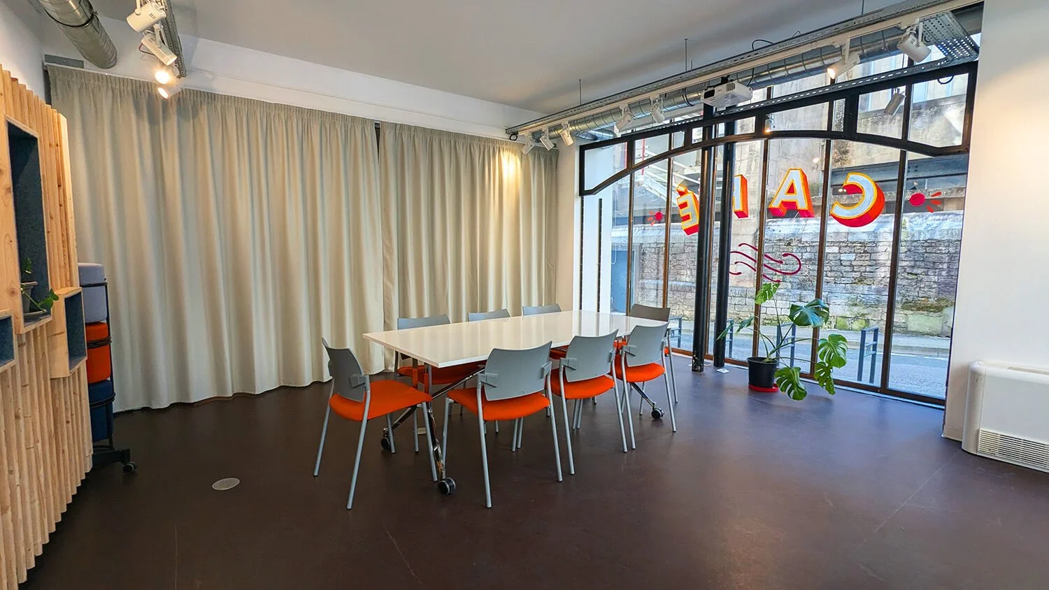 Une salle de réunion avec une table blanche entourée de neuf chaises à assise orange, un grand panneau de verre laissant entrer la lumière naturelle, des plantes vertes, des rideaux blancs, et une enseigne lumineuse rouge avec le mot 'Café' inversé en extérieur.