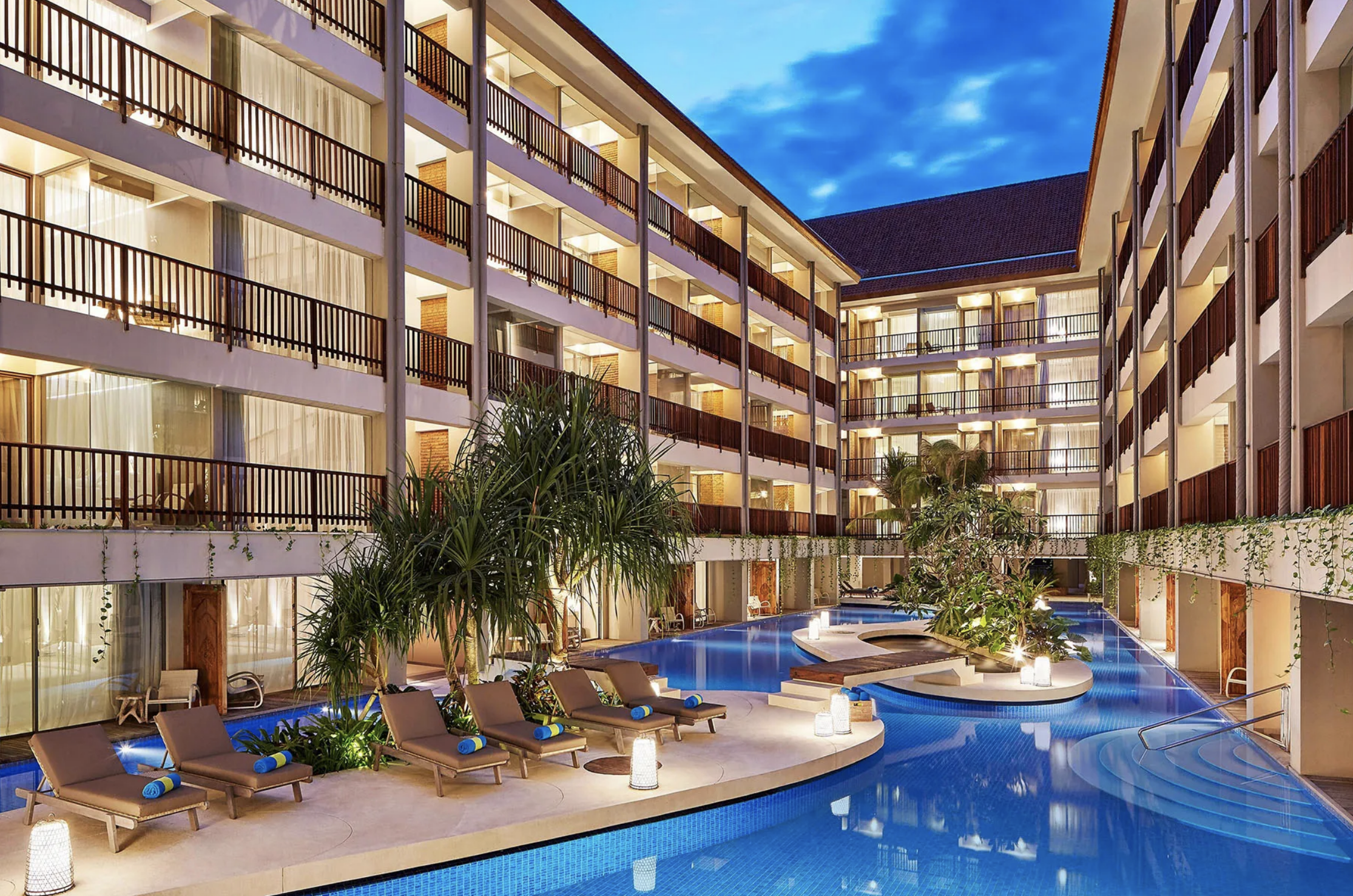 An evening view of a hotel courtyard with a swimming pool, lounge chairs with rolled towels, and an illuminated multi-story building with balconies, surrounded by trees and plants. The sky is dark blue with some clouds.
