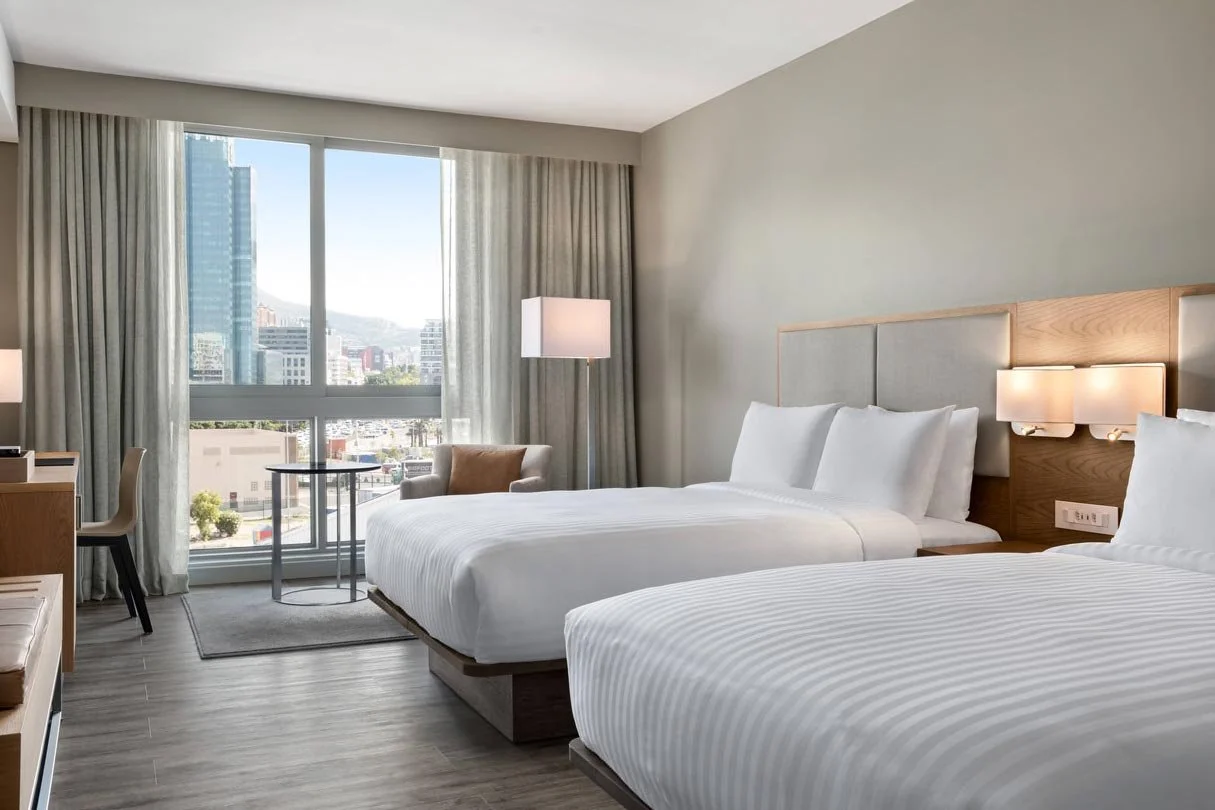 Hotel room with two beds, a large window showing city buildings, a small table with a chair and a cozy armchair, and modern lighting fixtures.