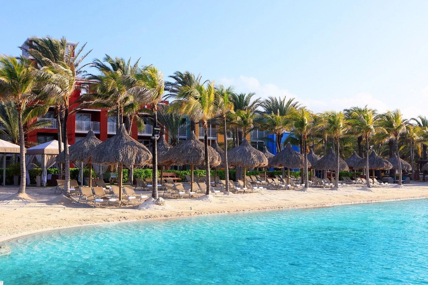 Beach scene with clear blue water, sandy shore, sun loungers, straw umbrellas, and palm trees with colorful buildings in the background.