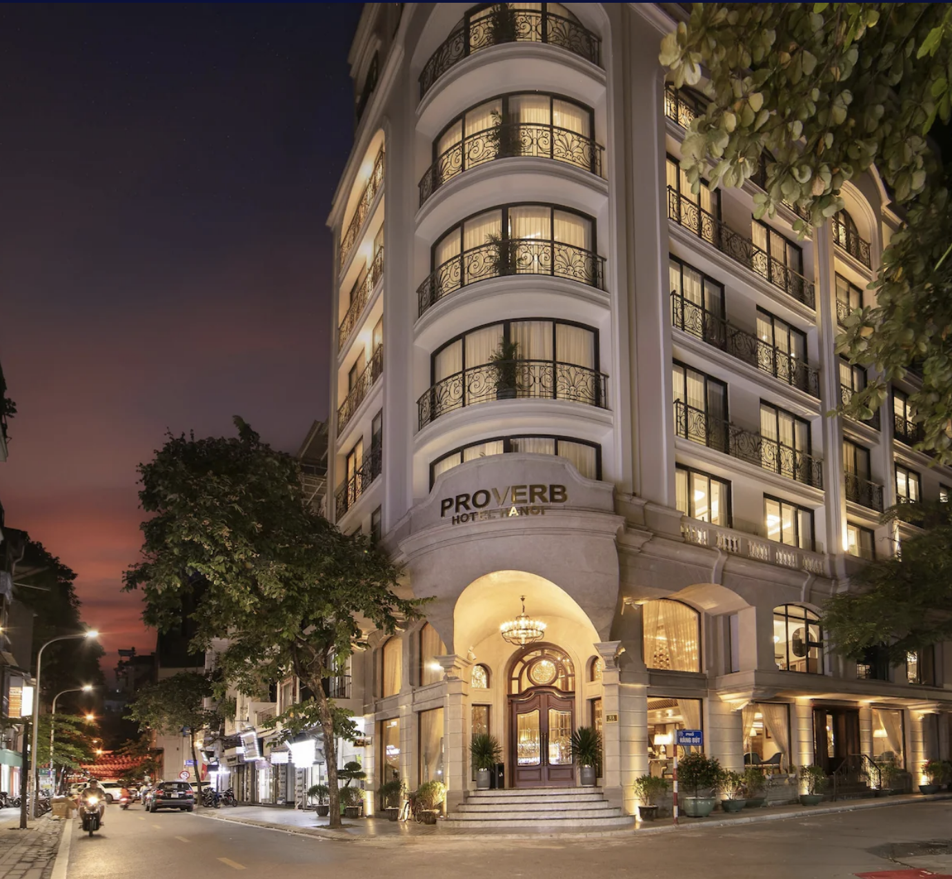 A multi-story hotel with a curved white facade and black wrought-iron balconies, illuminated at night with warm lighting from inside. The hotel is named 'Proverb Hotel & Spa' and is located on a busy street with trees, streetlights, and cars, against a twilight sky.