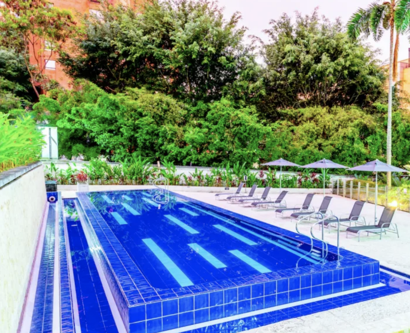 Outdoor swimming pool surrounded by lounge chairs and umbrellas, with lush green trees in the background.