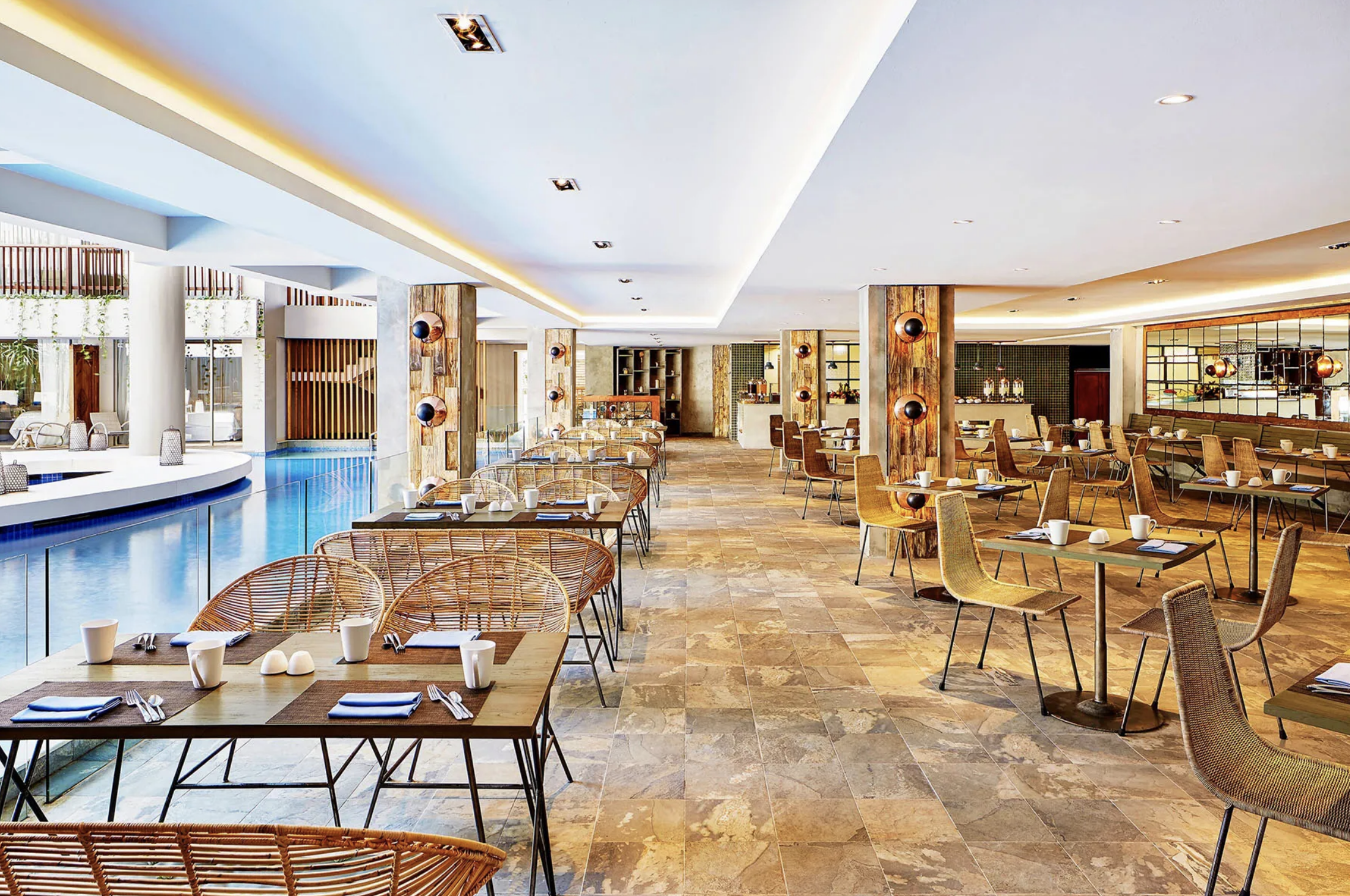 Spacious restaurant with tables set for dining, with cups, plates, and utensils, featuring a modern interior design with wooden accents, tiled flooring, and large windows.