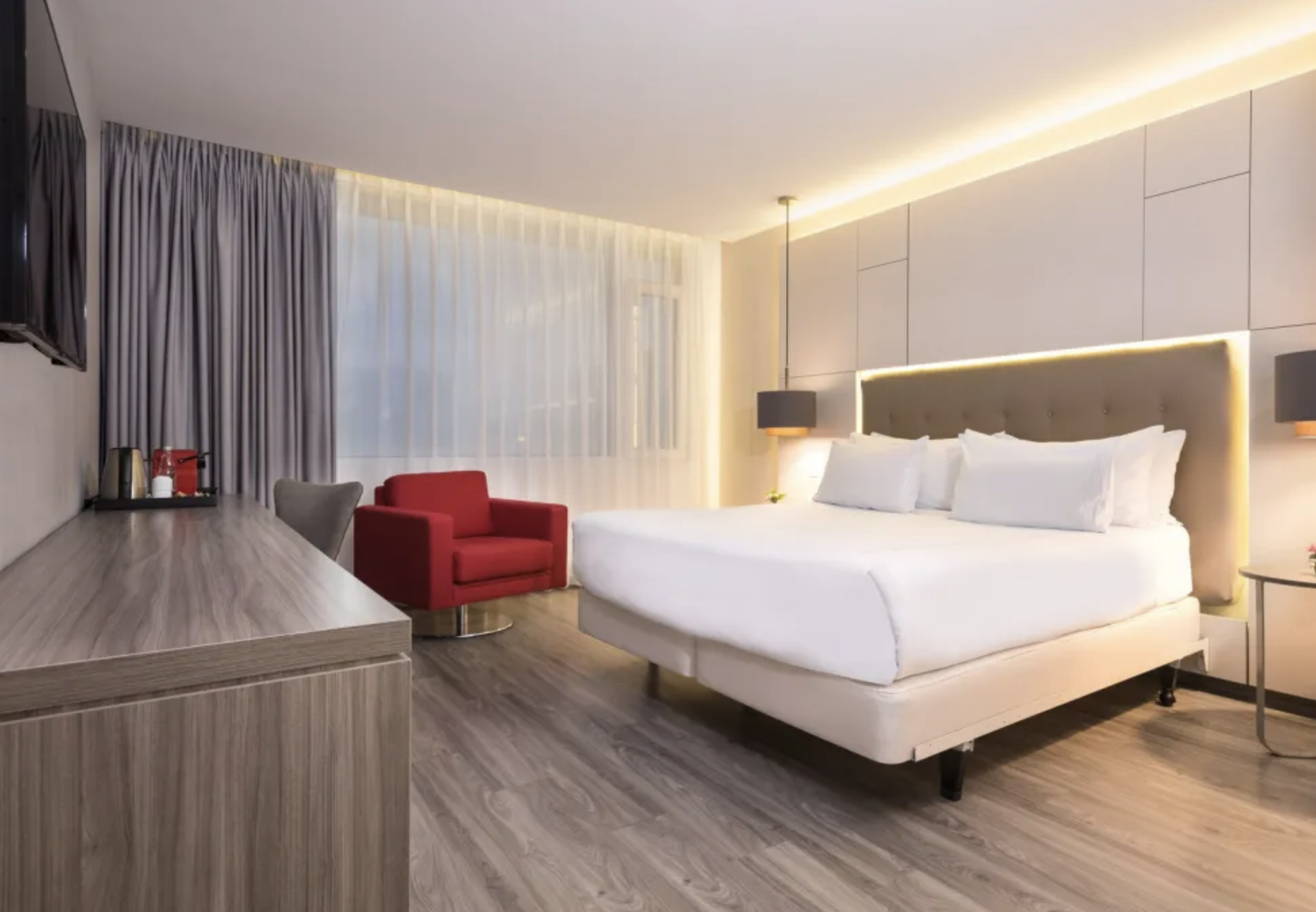 Modern hotel room with a large white bed, a red armchair, a gray chair, a wooden desk with a coffee station, large windows with sheer curtains, and minimalist lighting fixtures.