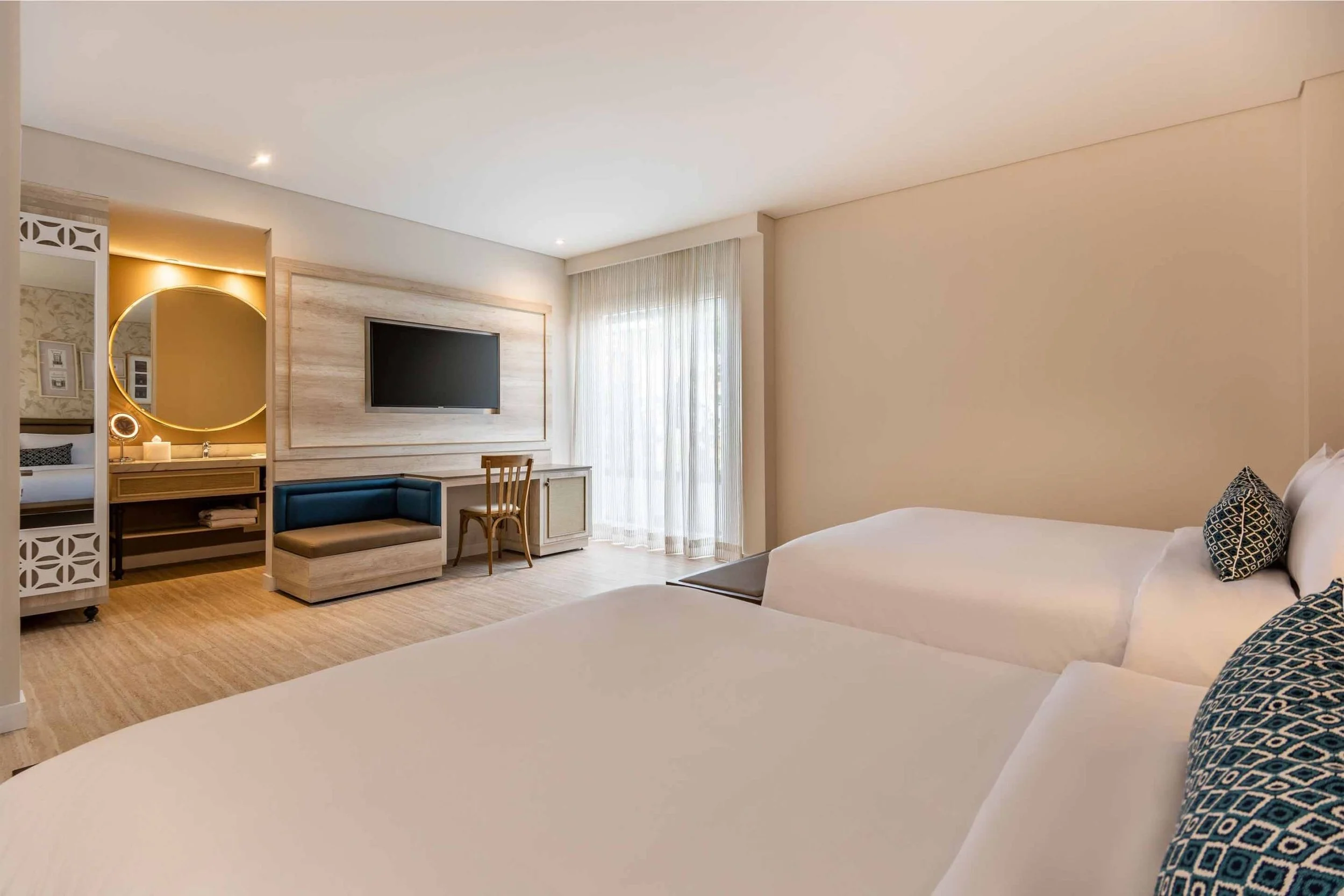 A hotel room with two beds, a television on the wall, a small seating area, a large window with sheer curtains, and a bathroom with a round mirror and sink visible through an open doorway.