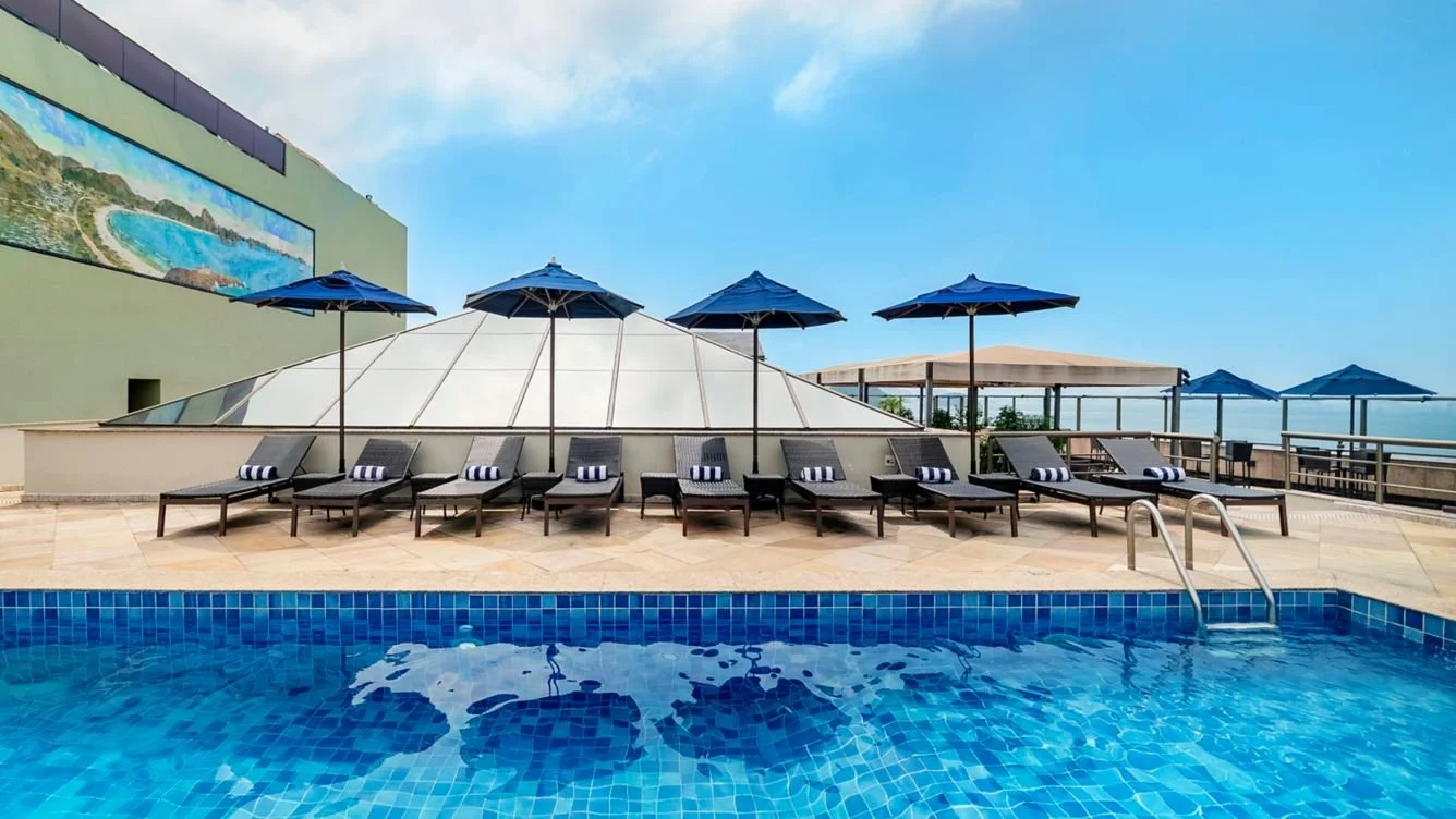 Swimming pool with blue tile mosaic, lined with black lounge chairs and blue umbrellas, overlooking a rooftop view with a building and sea in the background.