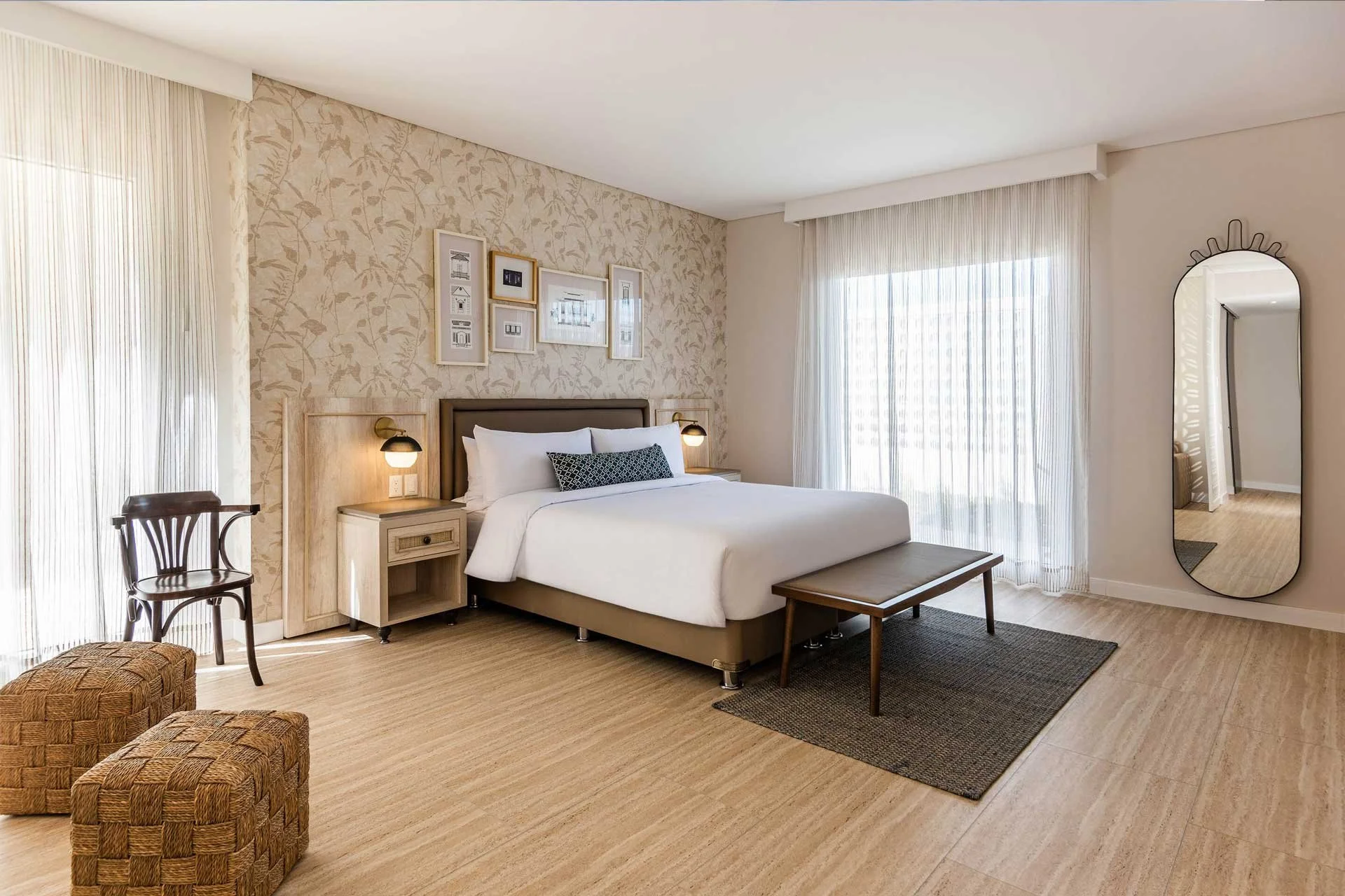 A bedroom with a large bed, white bedding, and decorative pillows. Light wooden furniture, including nightstands and a bench. Framed pictures above the bed, a mirror on the wall, and sheer curtains covering windows. Two woven ottomans and a patterned chair are visible.