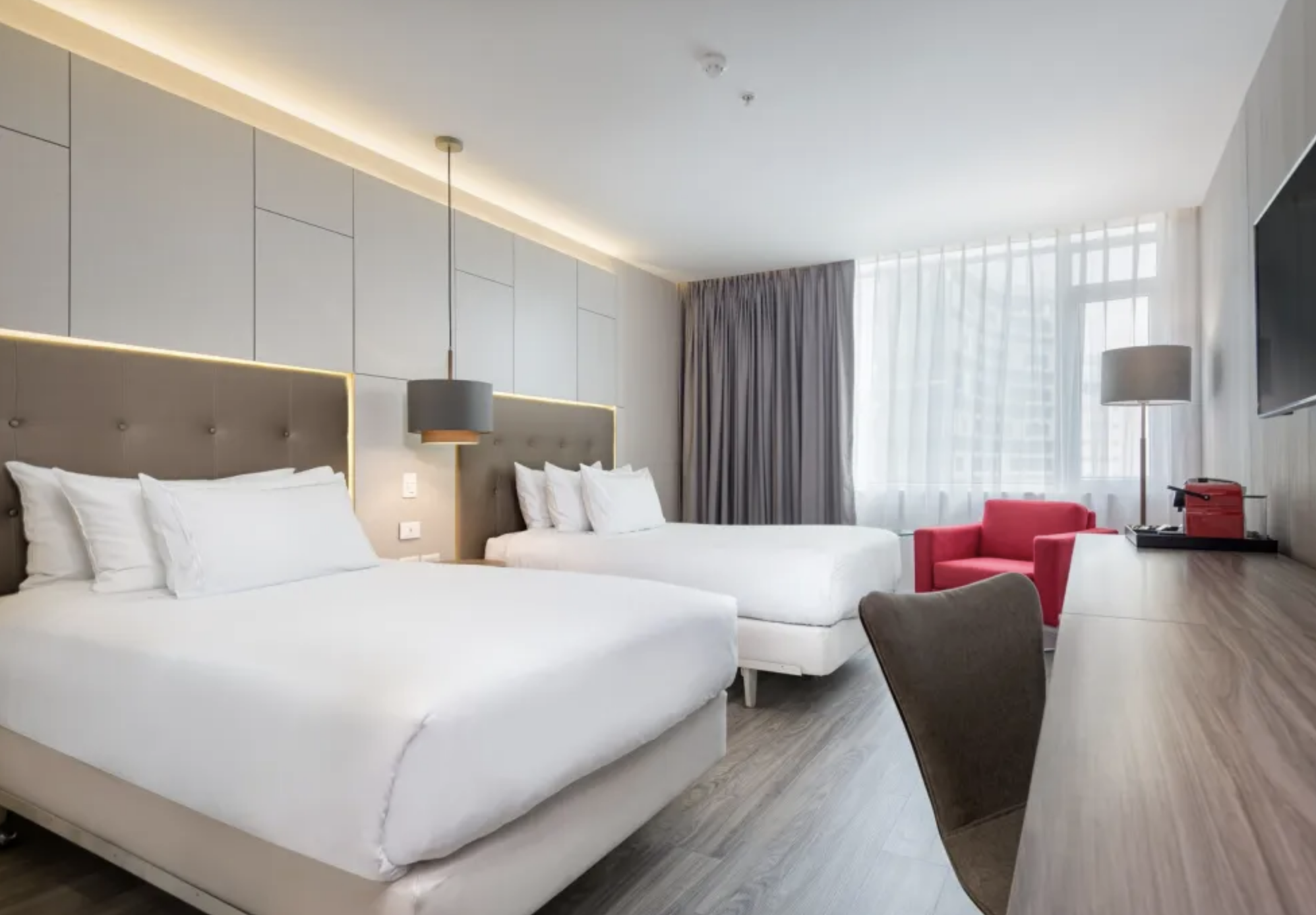 A modern hotel room with two white beds, beige headboards, bedside lamps, a red armchair, a large window with curtains, and a wall-mounted flat-screen TV.