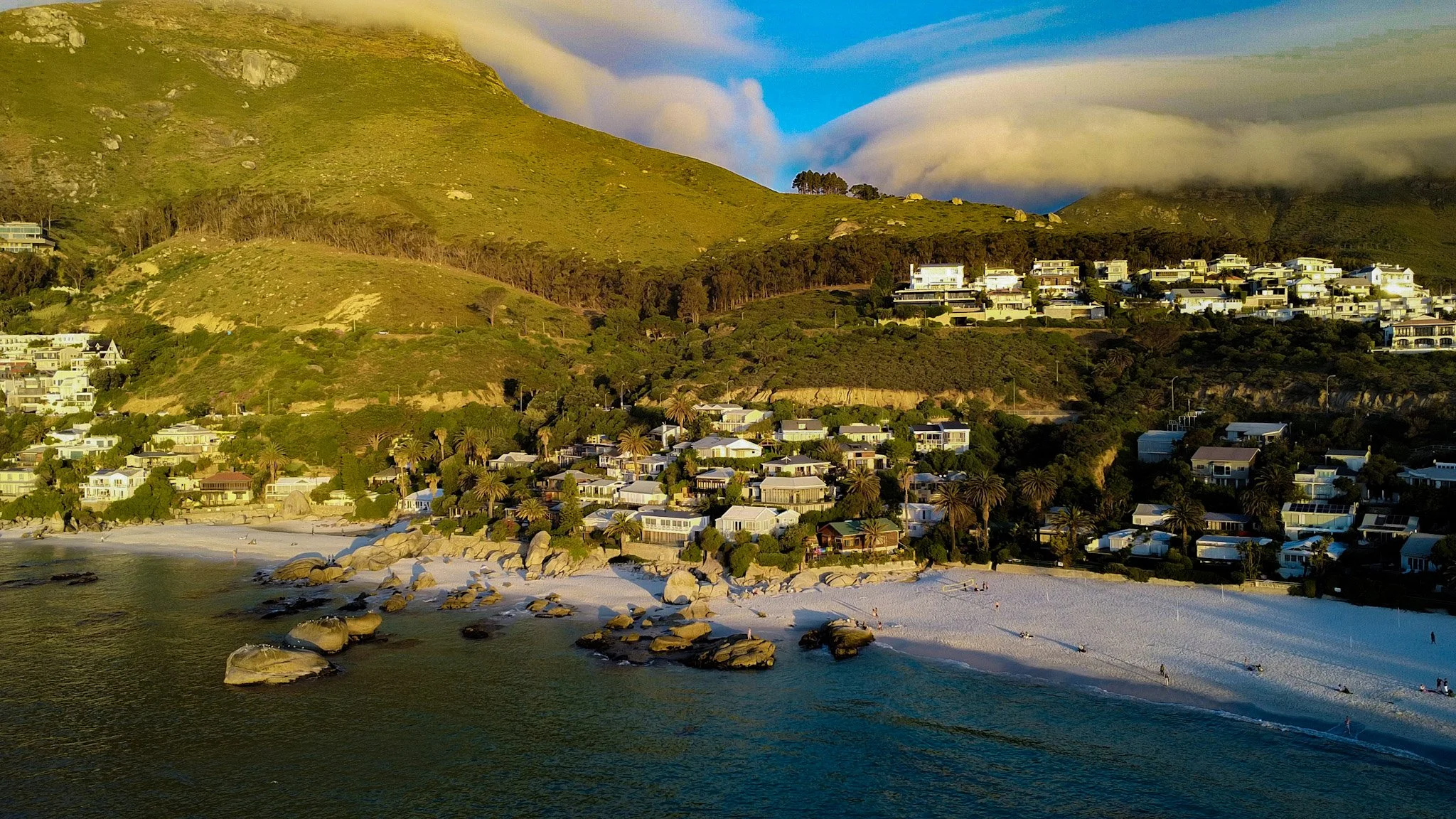 A coastal landscape with a beach in the foreground, rocks along the shoreline, and a hillside with numerous houses and trees in the background. The sky is partly cloudy, and the overall scene is illuminated by warm sunlight.