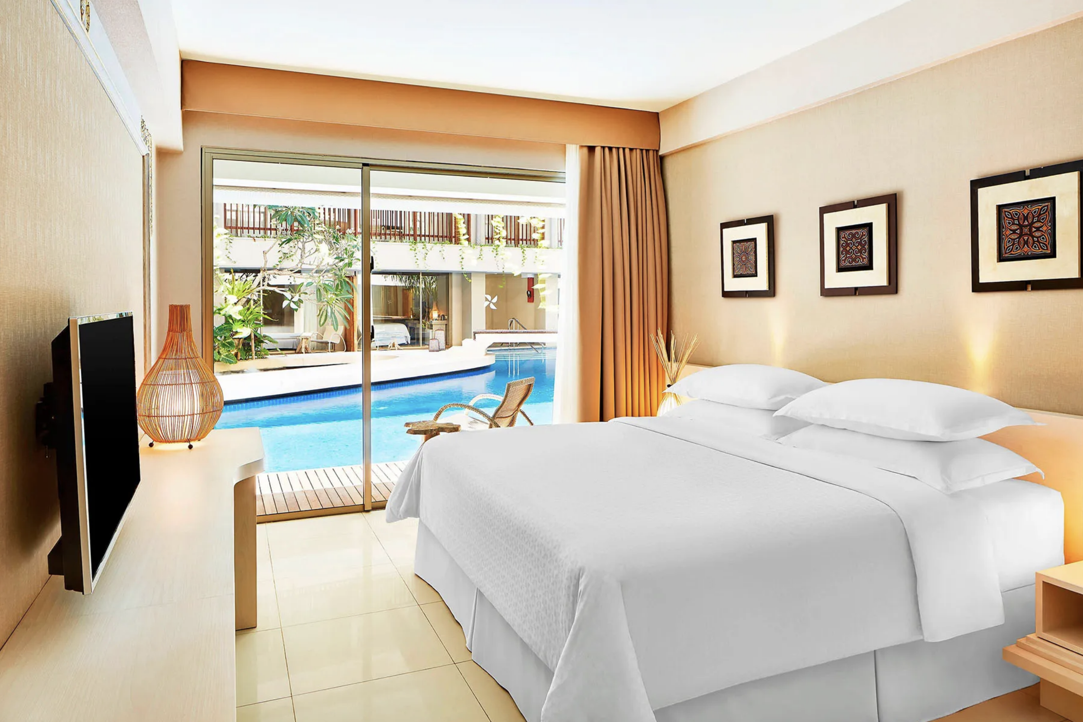 A hotel room with a large bed, wooden nightstand, and decor. There are three framed artworks on the wall and a sliding glass door leading to a balcony with a swimming pool outside.