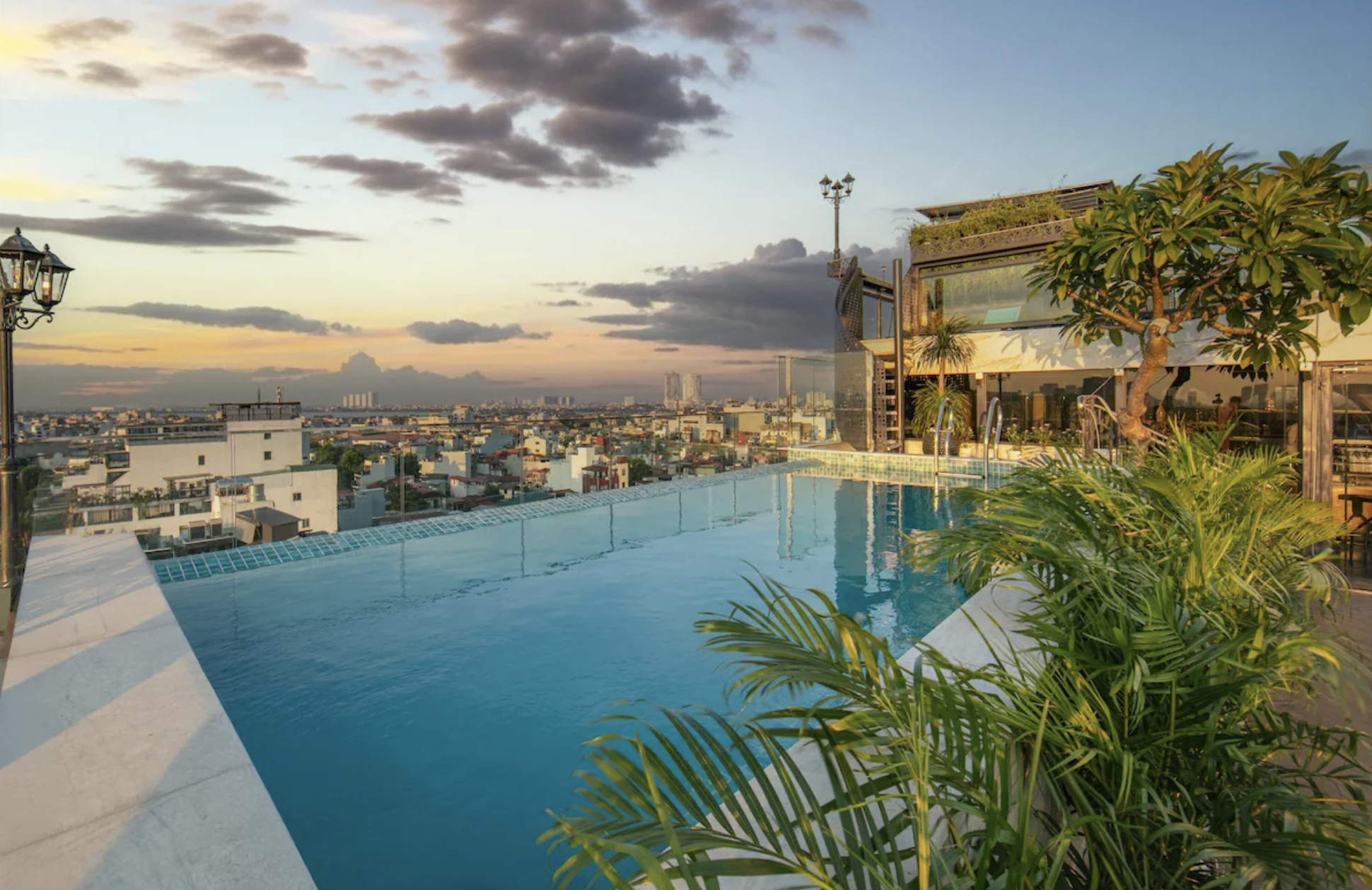 A rooftop infinity pool overlooking a city skyline during sunset, with plants and modern buildings around.