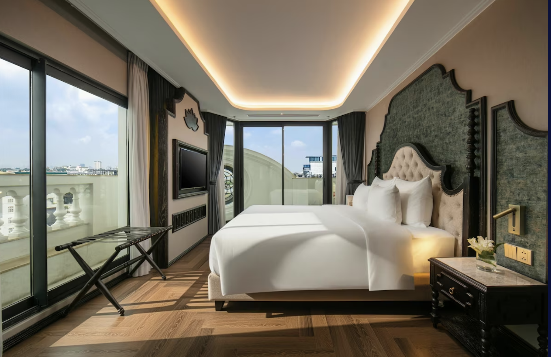 A modern hotel room with a large bed, dark headboard, and large windows with view of city buildings and the sky, featuring natural and warm lighting.