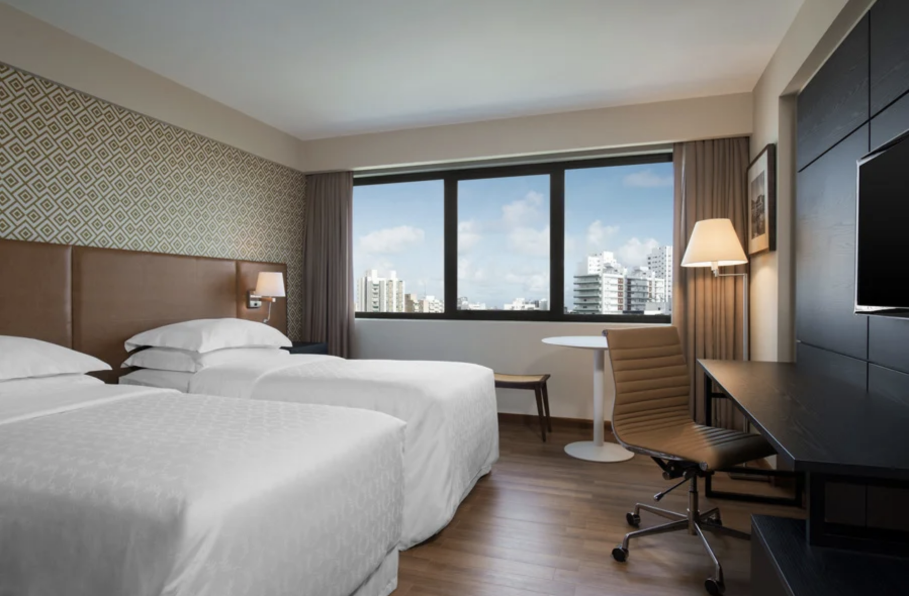 Interior of a modern hotel room with two beds, a large window showing city buildings, a small round table, a beige chair, a lamp, and a flat-screen TV on a black wooden stand.