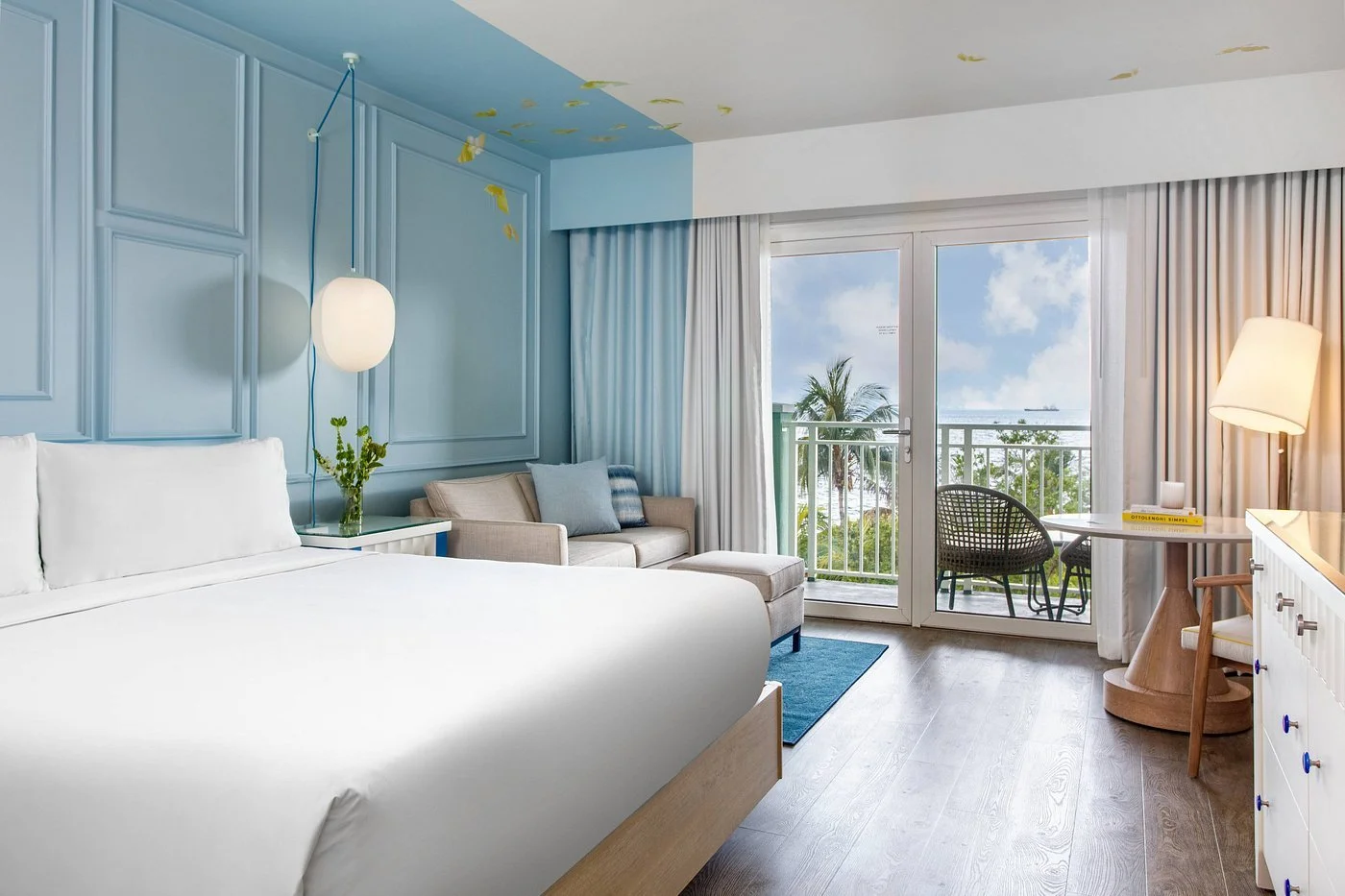 A bright hotel room with a large white bed, a beige sofa, and a glass sliding door opening to a balcony with ocean view, palm trees, and a ship in the distance.
