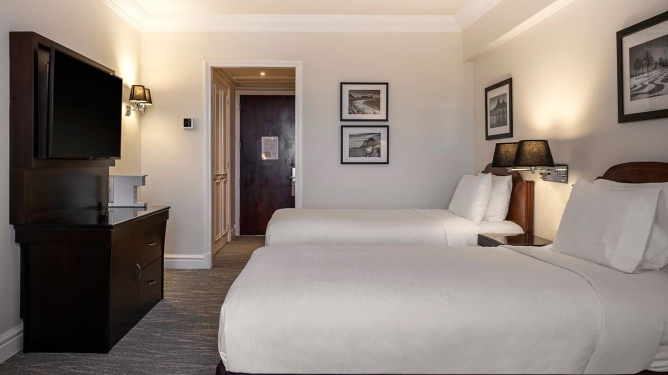 Hotel room with two white beds, dark wooden headboards, bedside lamps, framed black-and-white art, a dark wooden dresser with a television, carpeted floor, and entrance door.