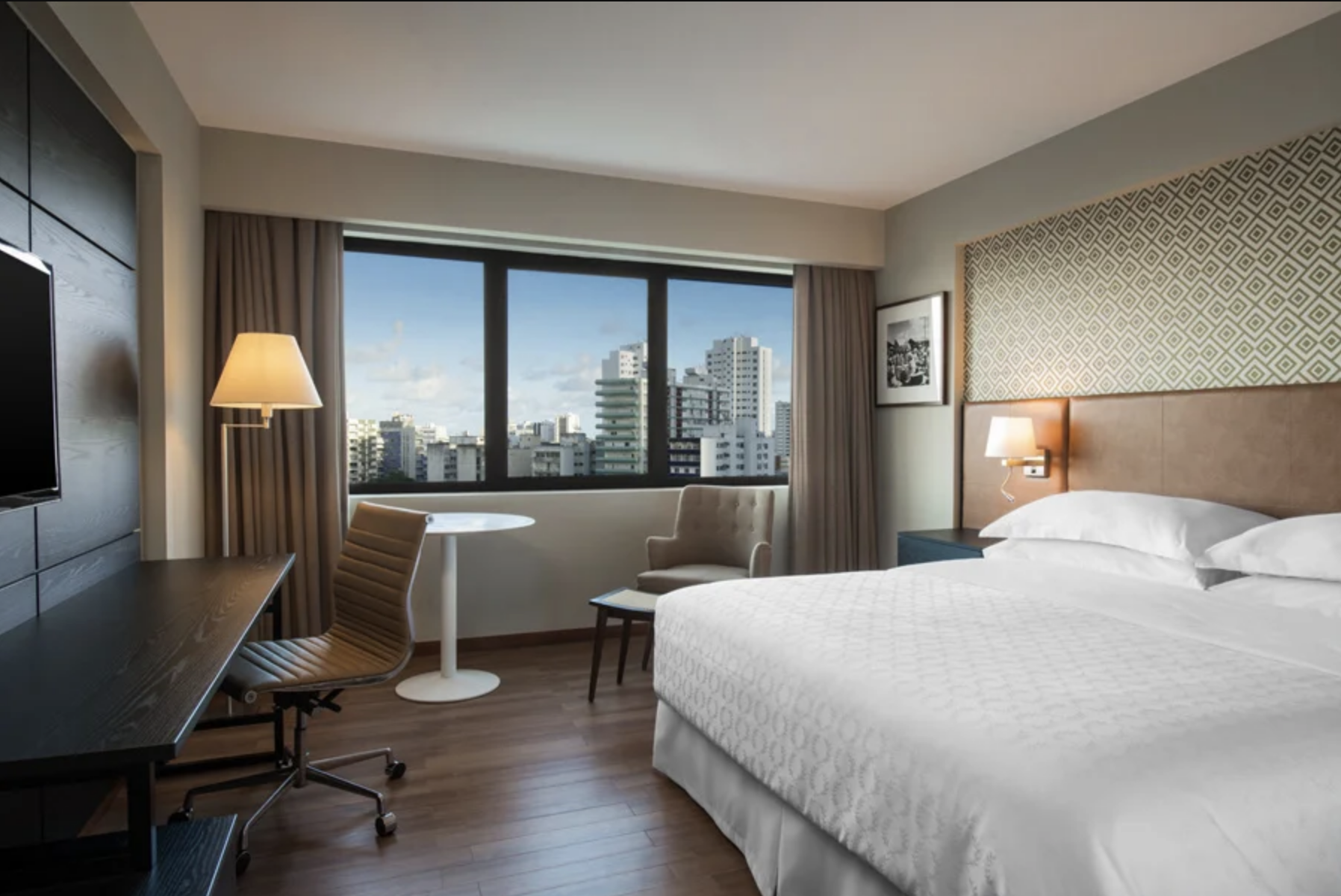 A modern hotel room with a large bed, a window with city view, a desk with a chair, a small table with an armchair, and wall decor.