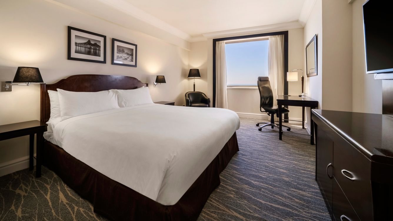Hotel room with a large bed, built-in lamps, black and white framed artwork, a window with a sea view, a desk with a chair, and a television.