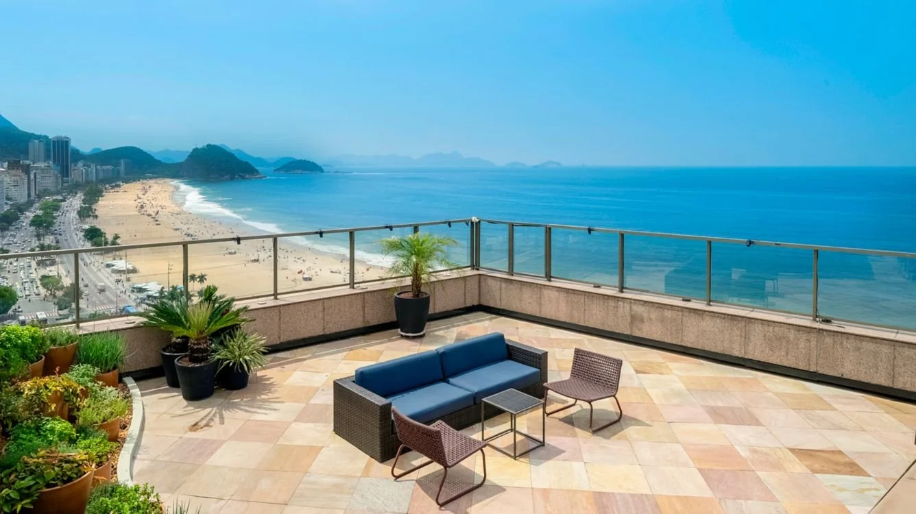 A rooftop terrace with outdoor seating including a sofa and two chairs, surrounded by potted plants, with a view of a beach, ocean, and cityscape in the distance.