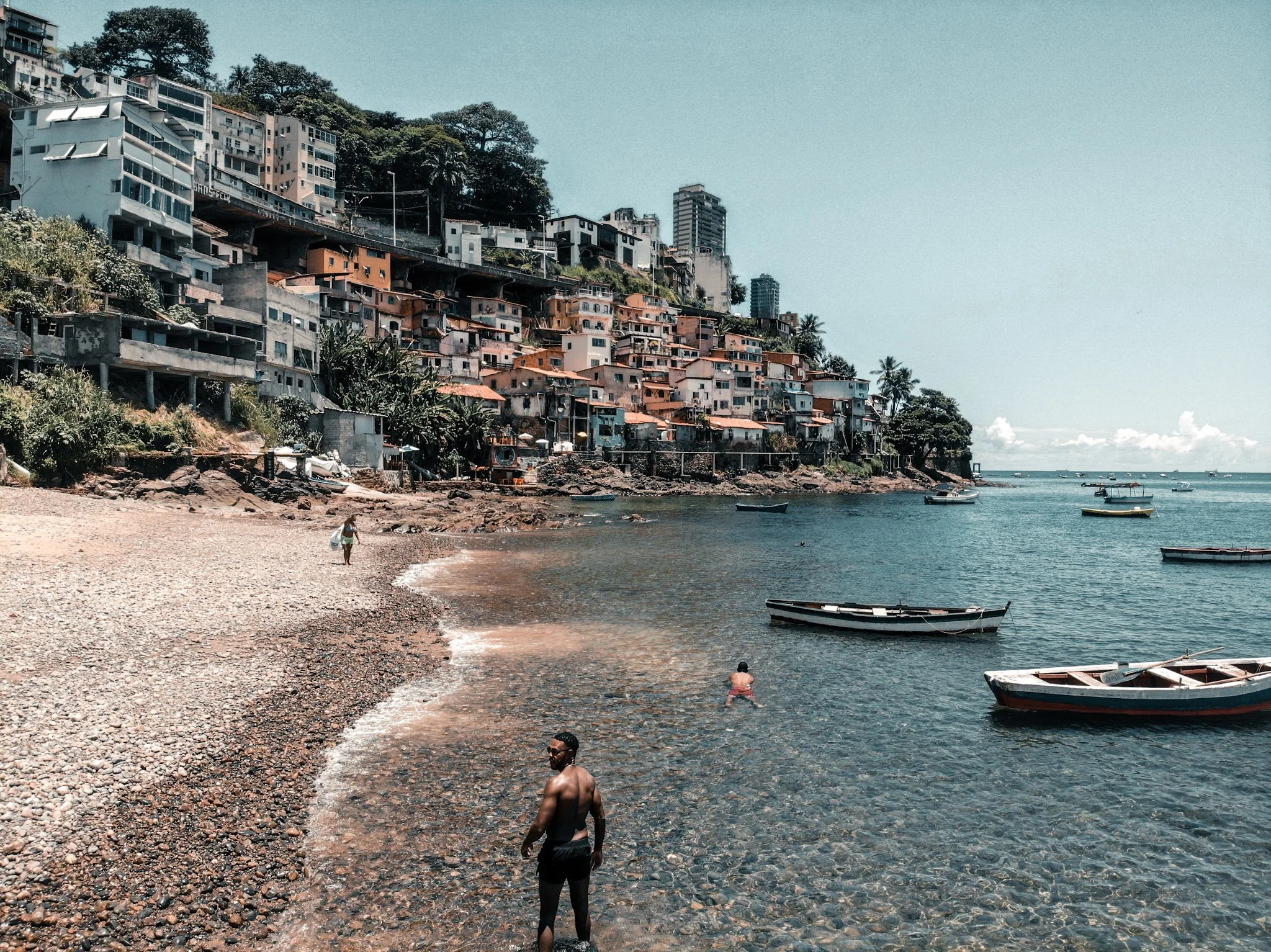 People relaxing and swimming at a rocky beach with colorful hillside houses and boats on the water, under a clear sky.