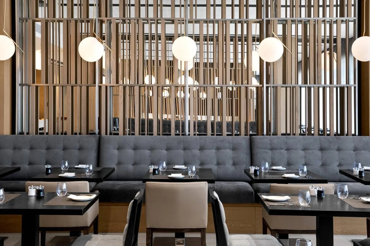 Interior of a modern restaurant with a bar seating area, wooden panel dividers with hanging spherical pendant lights, and tables set with glasses, plates, and utensils.