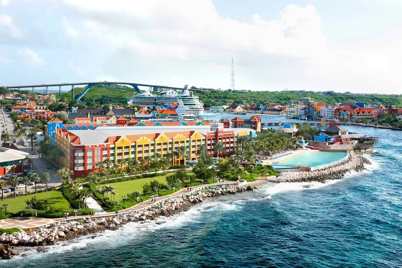 Colorful beachfront buildings, a large cruise ship, a bridge in the distance, and a water pool by the shoreline.